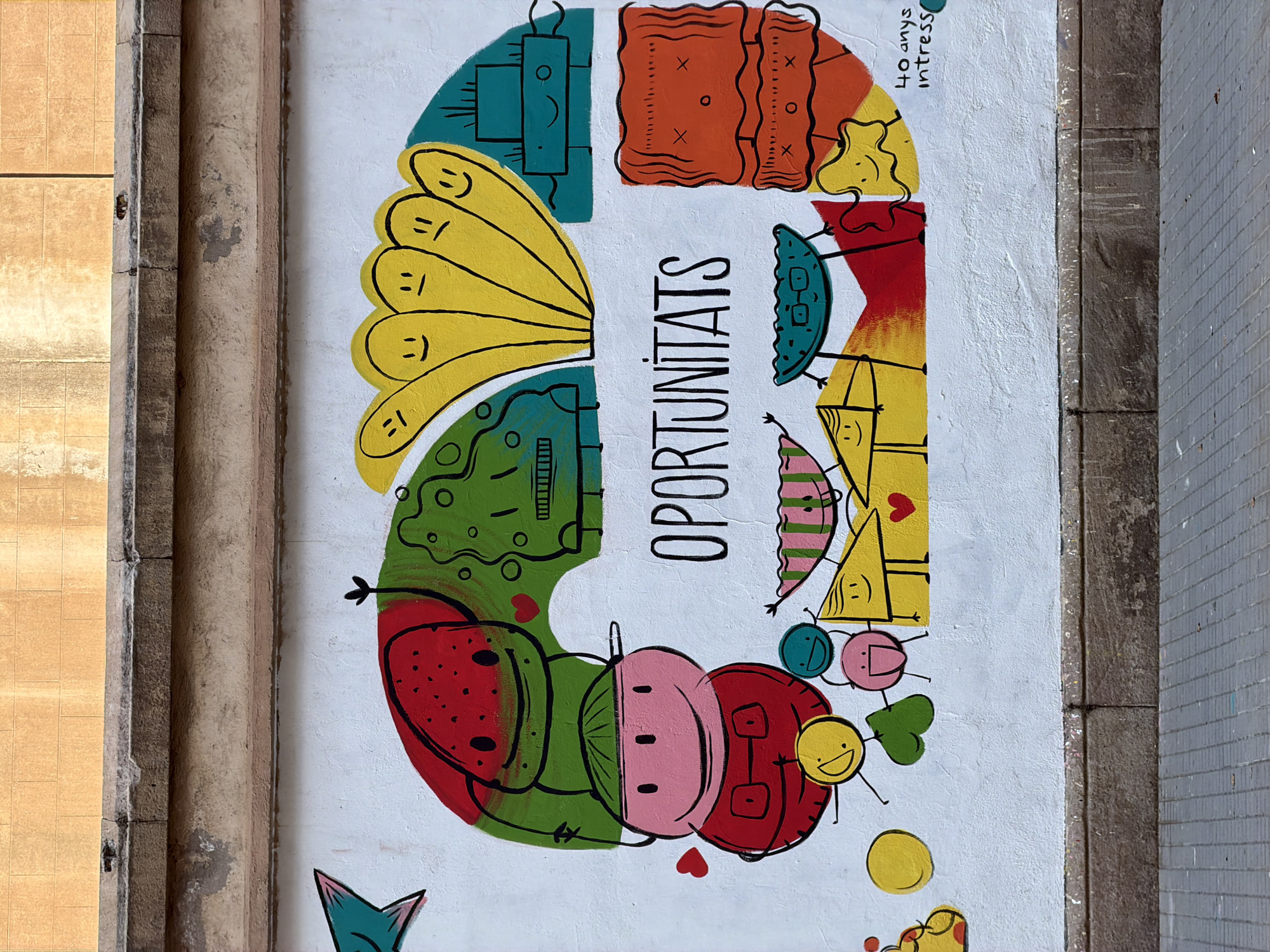 This is a photograph of a mural painted on a white textured wall, located in Barcelona, Spain. The mural features a colorful, cartoonish illustration of various abstract figures with smiling faces, arranged in a semi-circular arc. The figures are depicted with simple shapes and bold black outlines. The word "OPORTUNITATS" is prominently displayed in the center of the mural in large, black, block letters.
On the left side of the mural, a large red figure resembling a watermelon with black seeds and a smile is positioned above a pink, round-faced character wearing a red shirt and a green hat. Below these figures are several smaller, brightly colored abstract shapes, some with faces.
The upper portion of the mural shows a cluster of five yellow, finger-like figures with smiley faces and distinct expressions. To the right of these yellow figures is a teal rectangular figure with a smile and eyes.
The right side of the mural includes a tall, orange rectangular figure with wavy lines and "x" shapes, and a yellow, star-like figure with a smile. In the bottom right corner of the mural, handwritten text reads "40 anys intress".
The mural is painted above a concrete sidewalk with a pattern of gray square tiles. A stone ledge with a rough texture separates the mural from the sidewalk. The wall above the mural is painted in a warm, light orange color, with some texture and discoloration visible. The overall scene suggests an outdoor urban setting.