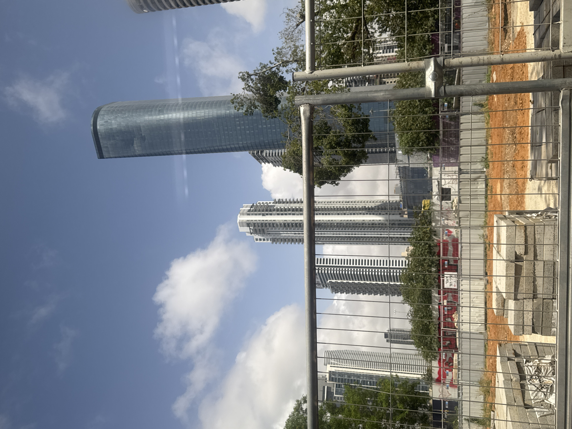 The image depicts an urban landscape, likely Tel Aviv as hinted, characterized by modern high-rise buildings and an active construction site in the foreground. The photograph is taken during daytime under a bright, clear blue sky with scattered white clouds.
In the immediate foreground, a metal construction fence, composed of vertical and horizontal bars forming a grid, occupies the lower and right portions of the frame. Behind this fence, a construction area is visible, featuring reddish-brown earth and stacks of grey concrete blocks or pavers. Some additional construction barriers and signs, including one red one with indistinct white Hebrew text, are present in the mid-distance of this site.
Beyond the construction site, several prominent skyscrapers dominate the view. On the left, a tall, sleek, cylindrical or oval-shaped glass building, which appears to be the Azrieli Sarona Tower, reflects the sky and surrounding environment. To its right, another high-rise building with a white facade and numerous balconies or horizontal elements stands out. Other less distinct tall buildings can be seen further in the background. Scattered green trees and foliage are visible, some reflected in the glass surfaces of the modern buildings.
The bright lighting suggests it is either mid-morning or mid-afternoon. No specific activity or people are clearly visible, implying a static scene of urban development. The overall impression is one of a bustling city undergoing growth and modernization, viewed from a pedestrian perspective near a development area.