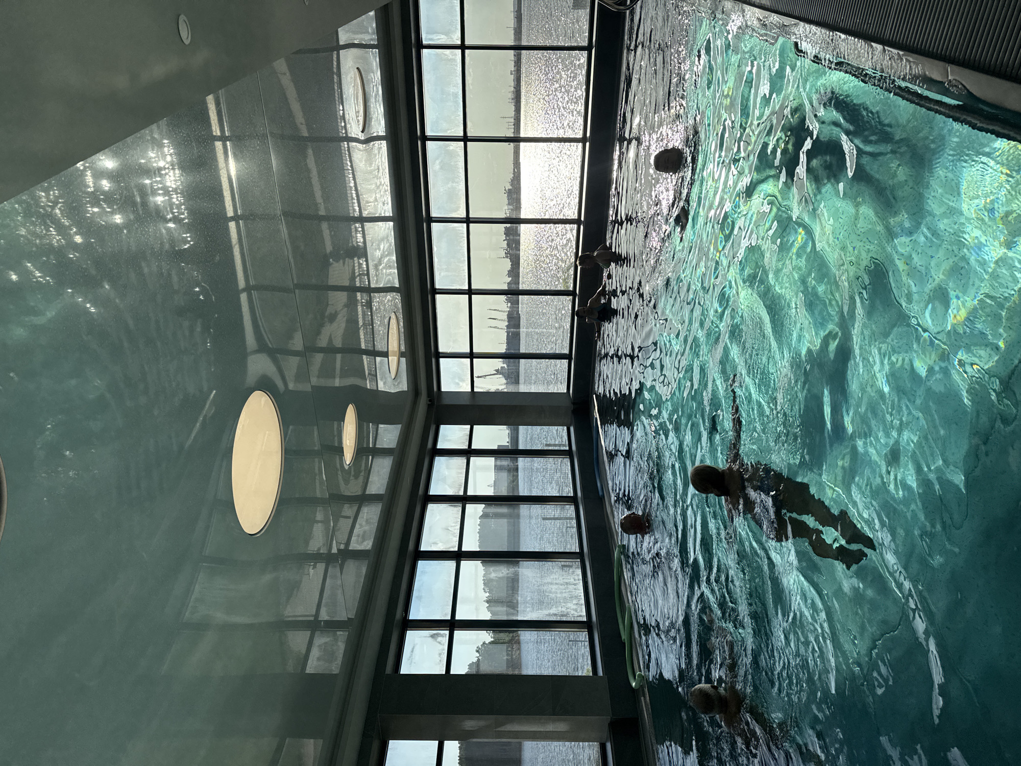 The image depicts an indoor swimming pool at a perpendicular angle, suggesting the photograph was taken either from above or from a tilted perspective. Several individuals are swimming in the turquoise water. A row of tall windows lines one side of the pool, providing a view of an outdoor body of water reflecting sunlight. The ceiling features round skylights. The scene takes place in the Aqua Chaikovsky swimming pool in Chaikovsky, Russia.