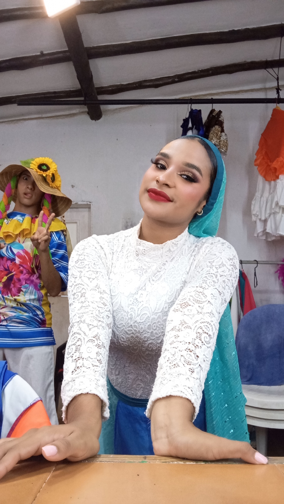 The image depicts an indoor scene in Porlamar, Venezuela. A young woman is positioned centrally and in the foreground, leaning forward with her hands resting on a light brown wooden surface. She looks directly at the viewer with a slight smile, prominent eye makeup, and red lipstick. She wears a white long-sleeved lace top, a teal head covering, and a gold hoop earring in her visible left ear. Her fingernails have pink polish. In the blurred background to the left, a second individual stands, wearing a straw hat adorned with sunflowers and colorful braids or ribbons, and a vibrant, multi-colored patterned shirt. This person is holding up two fingers in a 'peace sign' gesture and making a playful facial expression, with their tongue slightly extended. The background features light-colored walls, exposed dark wooden ceiling beams, and a bright rectangular overhead light fixture in the upper left. A metal rod serves as a clothes rack in the mid-background, from which various items of clothing or costume, including dark blue, leopard print, orange, and white ruffled fabrics, are hanging. A blurred white door or door frame is visible behind the second person, and a partial blue chair is visible in the lower right.