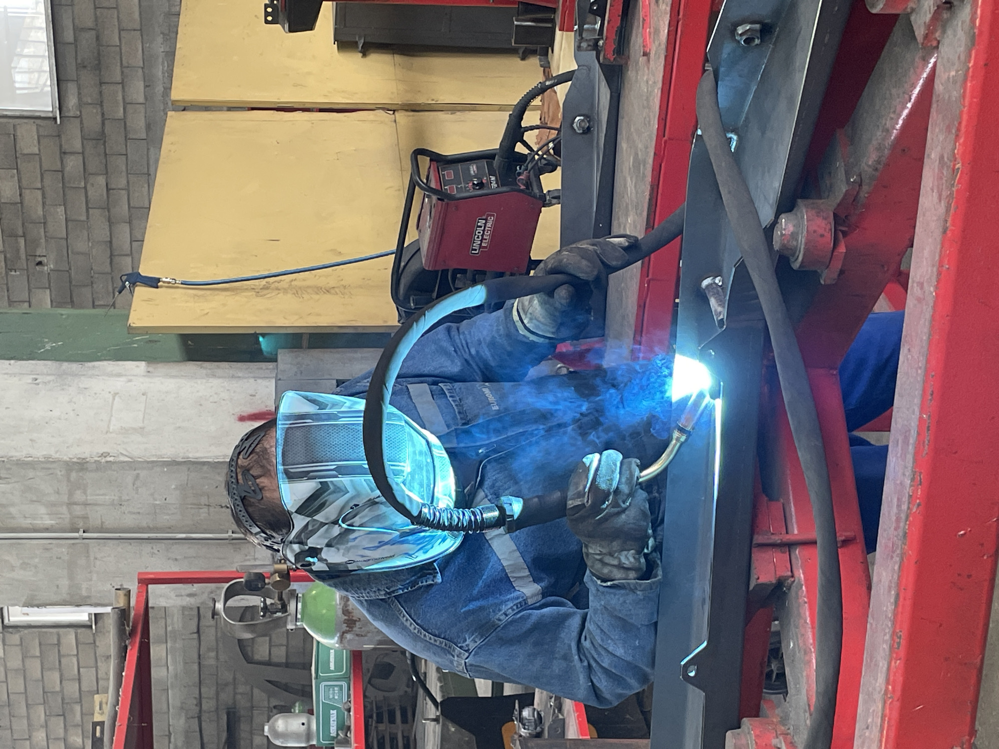 The image shows a person welding in what appears to be a workshop. The person is wearing a welding helmet, gloves, and a denim jacket. They are using a welding torch to join two pieces of metal together, with bright sparks visible. A Lincoln Electric welding machine is visible in the background, along with a red metal frame structure. A gas cylinder is also visible. The location of Čeluga, Montenegro, is not evident from the image alone.
