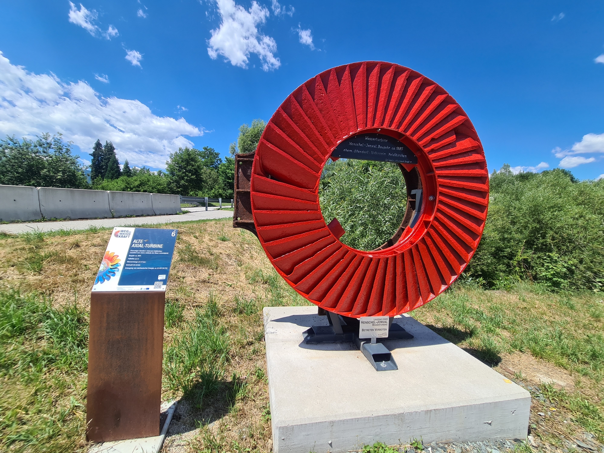 In Feldkirchen, Kärnten, Austria, a bright red, large axial turbine wheel, a historical artifact, is displayed outdoors on a concrete pedestal. The turbine, approximately 1.5 meters in diameter, has numerous fins radiating from its center and is mounted on a dark metal support structure. To the left of the turbine, a brown metal information stand displays a sign with text and graphics, including the title "WEG DER WASSERKRAFT" and "ALTE AXIAL-TURBINE" along with technical specifications and historical details. The turbine is situated in a grassy area bordered by a concrete barrier and lush green vegetation. The sky above is a vibrant blue with scattered white clouds. A small black plaque is visible on the turbine's base, bearing German text.