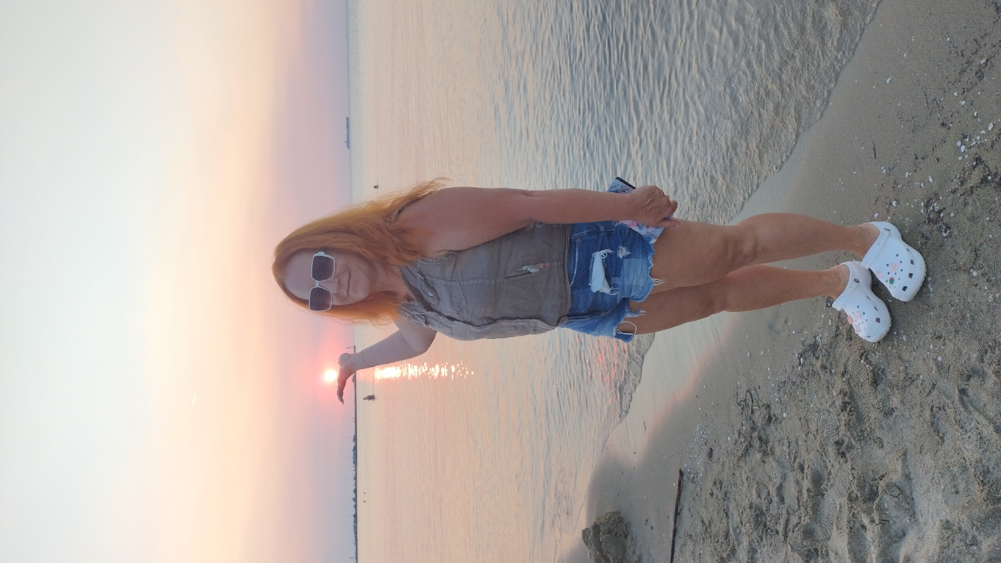 A woman with reddish-orange hair, wearing white-rimmed sunglasses, a tan sleeveless vest, and distressed denim shorts, stands on a sandy beach in Neoi Epivates, Greece, at sunset. She holds her left hand up, appearing to cup the setting sun. The sun casts a golden reflection on the calm sea. A dark object, possibly a phone, is held in her right hand. White Croc-style shoes with floral embellishments are on her feet. The background shows a hazy horizon with a faint silhouette of a ship and a pier with a few distant figures. The sand is scattered with small shells.