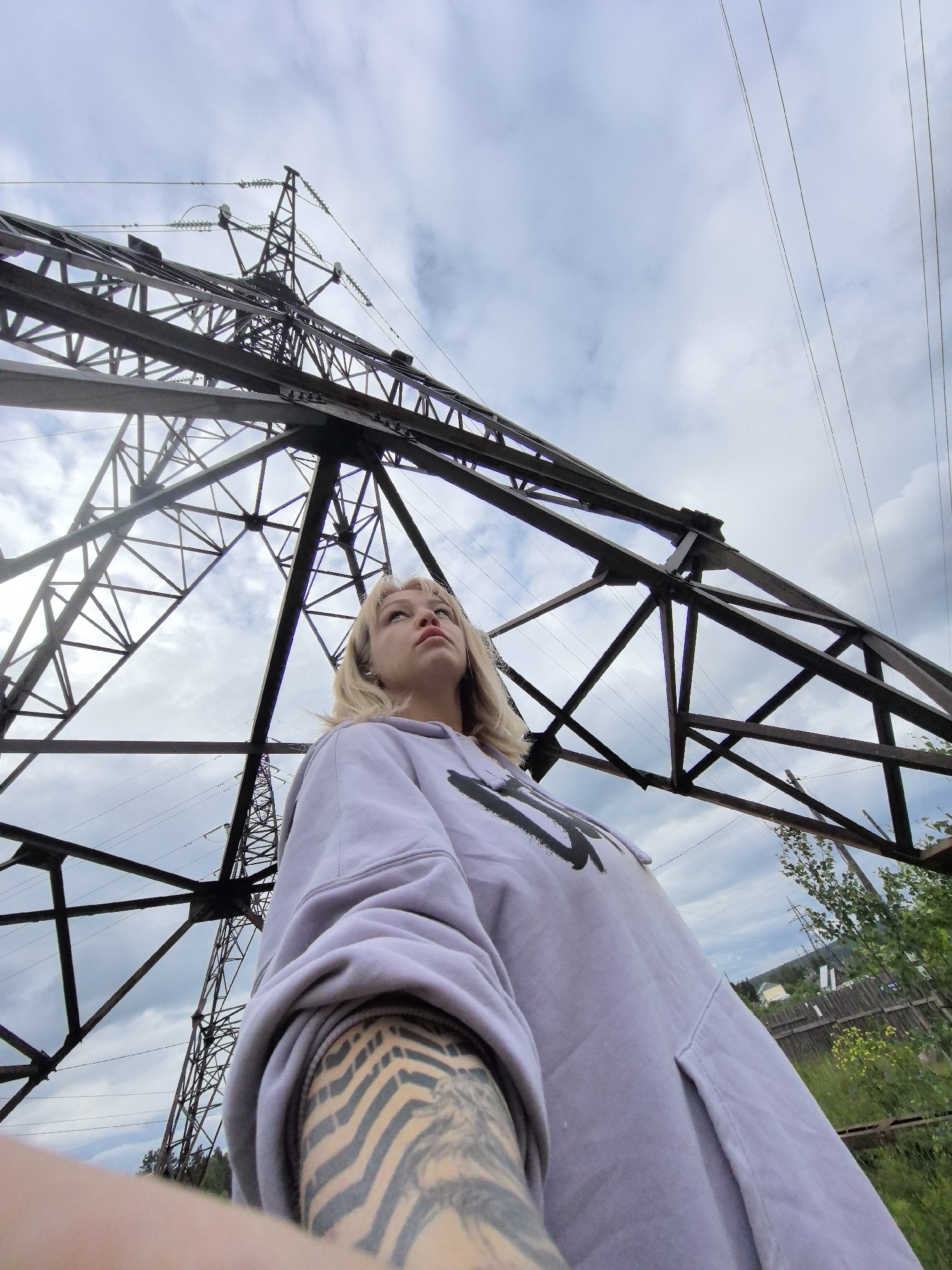 The image is a low-angle selfie taken by a young woman in Noviy, Russia, positioned directly beneath a massive metal electricity pylon. The woman, with blonde hair, is looking upwards and to her right, towards the vast structure and the sky. She wears a light purple, oversized hoodie with a dark, indistinct design on the chest. Her left arm is prominent in the foreground, revealing a sleeve tattoo featuring a striking striped pattern, possibly tiger stripes, along with other design elements.
The intricate, geometric lattice work of the electricity pylon dominates the upper and left portions of the frame, highlighting its imposing scale. Power lines stretch across the cloudy sky, which shows hints of blue. In the lower right and background, green foliage, including trees, bushes, and grass, suggests a natural or semi-rural setting. Distant houses or buildings, partially obscured by fences, are visible through the vegetation on the right. The lighting indicates it is daytime, under an overcast sky, creating soft and even illumination. The woman's expression is one of contemplation or curiosity as she gazes upwards. No legible text is visible in the image.