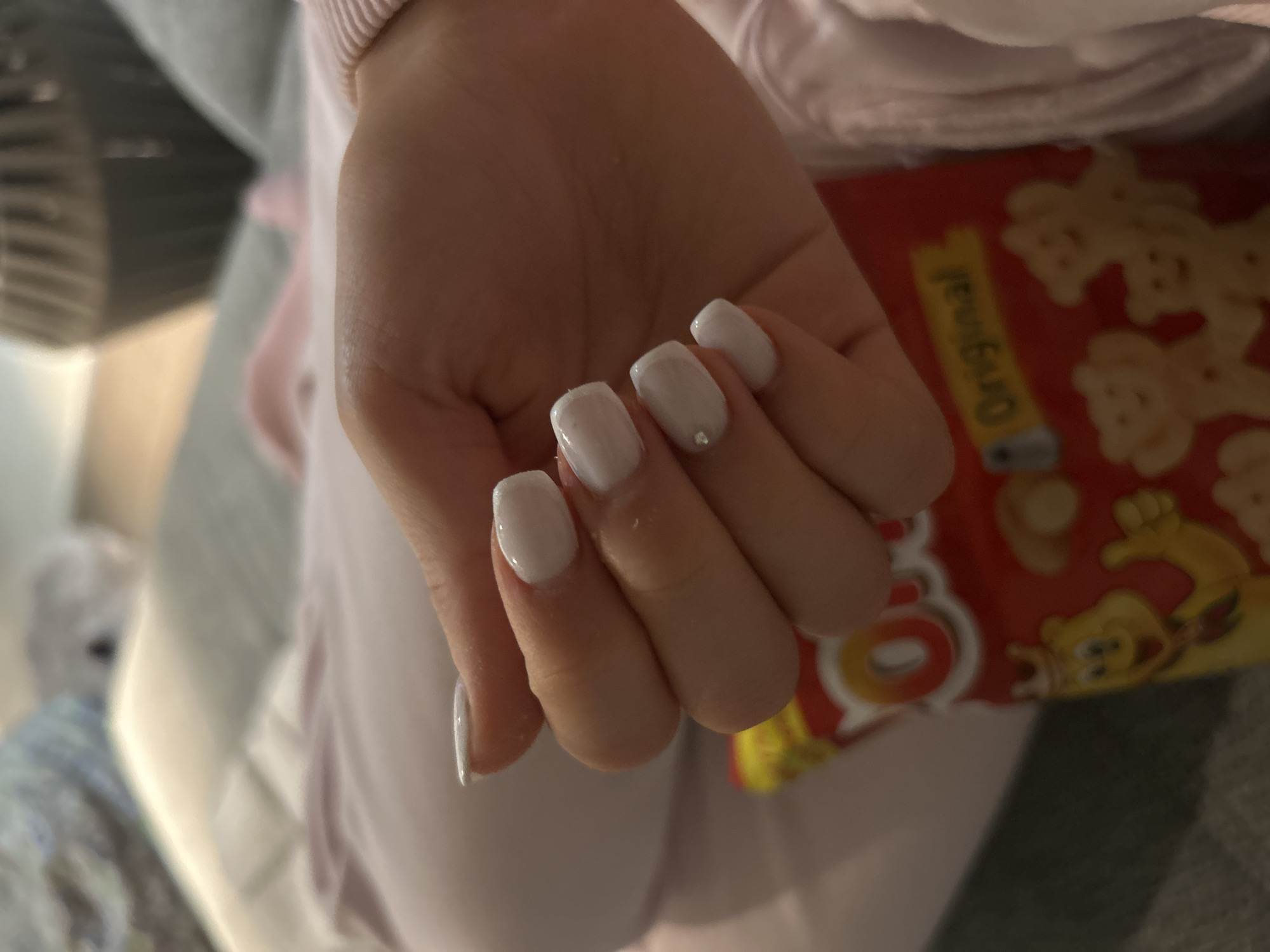 A close-up image shows a hand with white, square-shaped nails holding a red package of "Original" cookies. The hand is resting on a pink garment. In the background, part of a white fan and a blurry white object are visible.