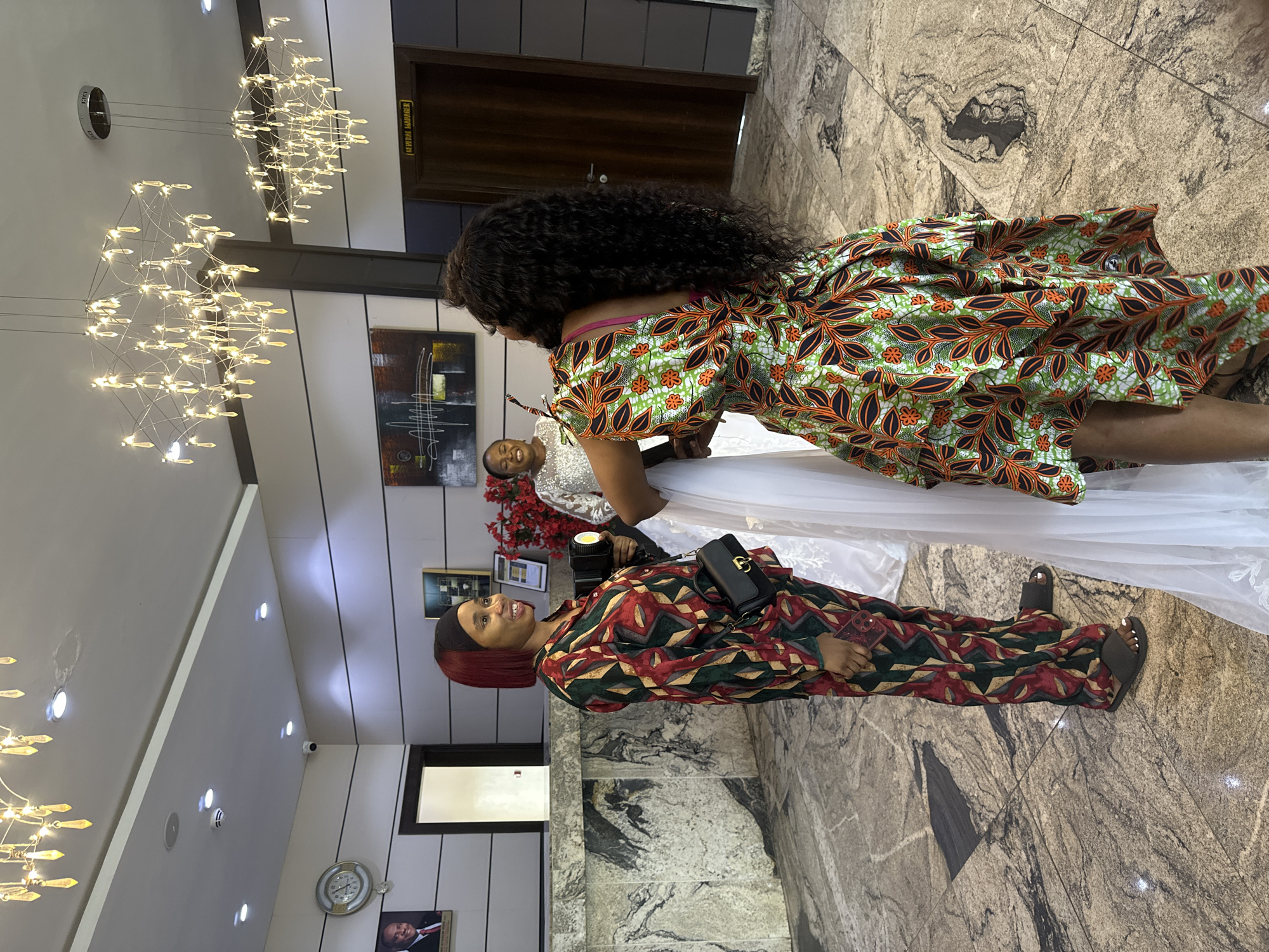 Three women are standing in an indoor, well-lit space that appears to be a hotel lobby in Abuja, Nigeria. The floor is tiled with a marbled pattern. Above, two modern chandeliers with multiple crystal-like accents illuminate the area.
On the left, a woman with short, dyed red hair and a black headscarf is wearing a vibrant, patterned pajama-style outfit in shades of red, green, and gold. She is holding a small black bag and what appears to be a portable light or camera attachment. She is looking to her right, smiling.
In the center, a woman in a white, shimmering dress is partially visible. She is smiling and looking forward.
On the right, a woman with long, dark, curly hair is wearing a colorful dress with a green base and a bold orange and black floral leaf pattern. She is facing away from the camera, holding onto the white dress.
In the background, a reception desk is visible on the left, with a framed portrait of a man in a suit hanging on the wall above it. To the right, a wooden door with "GENERAL MANAGER" signage is present. Red floral arrangements are also visible.