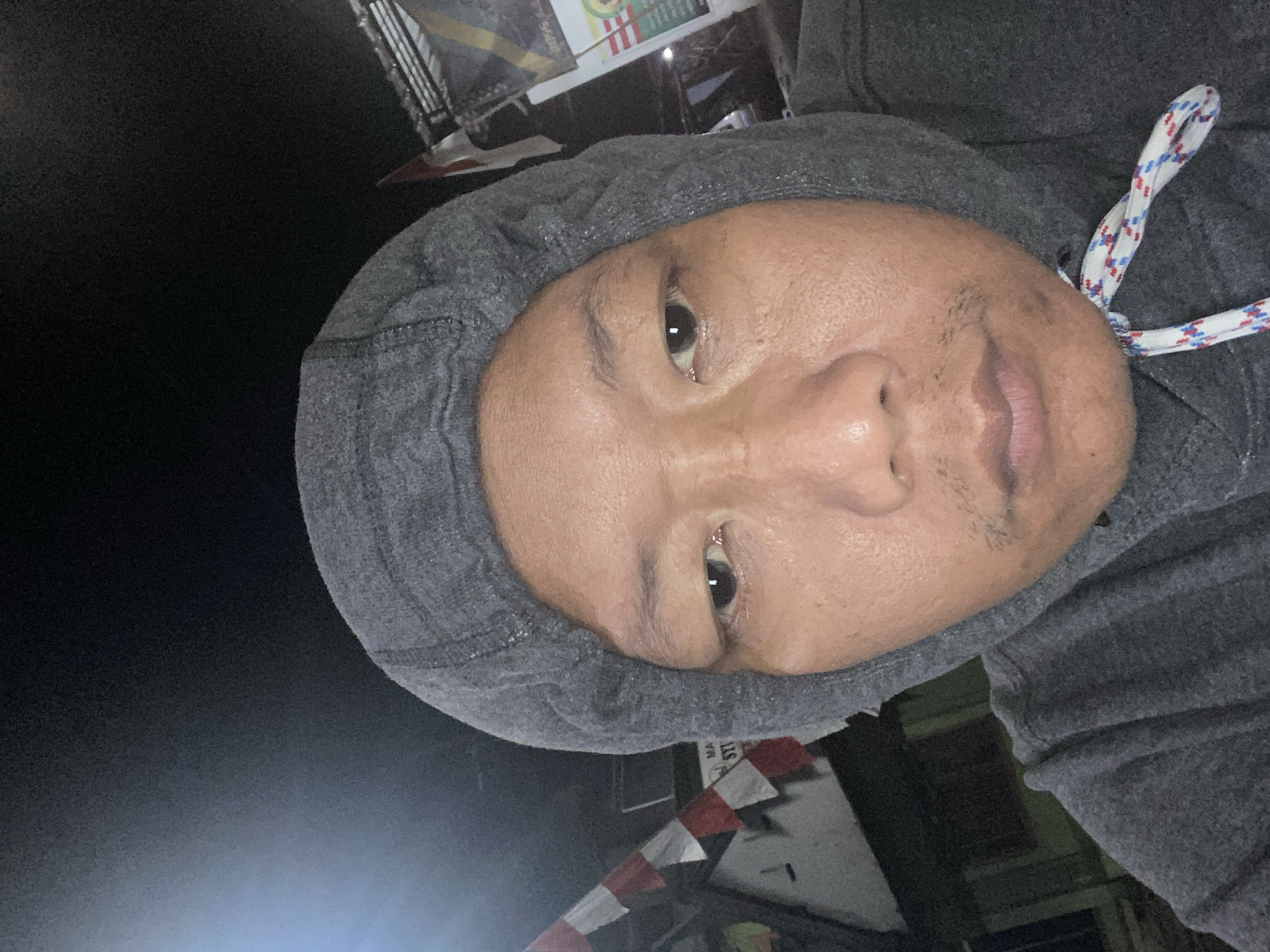 A close-up, selfie-style photograph shows a person wearing a grey hooded sweatshirt. The person's face is visible, with their eyes open and looking towards the camera. The background is dark, suggesting it is nighttime or a poorly lit area. To the left of the person, a flag with red and white stripes is partially visible, potentially indicating a location in Indonesia. Behind the person, there are hints of buildings and other objects, but they are indistinct due to the low light and focus on the subject. The location in Tulungagung, Indonesia, is not explicitly depicted but is inferred from the context provided.
