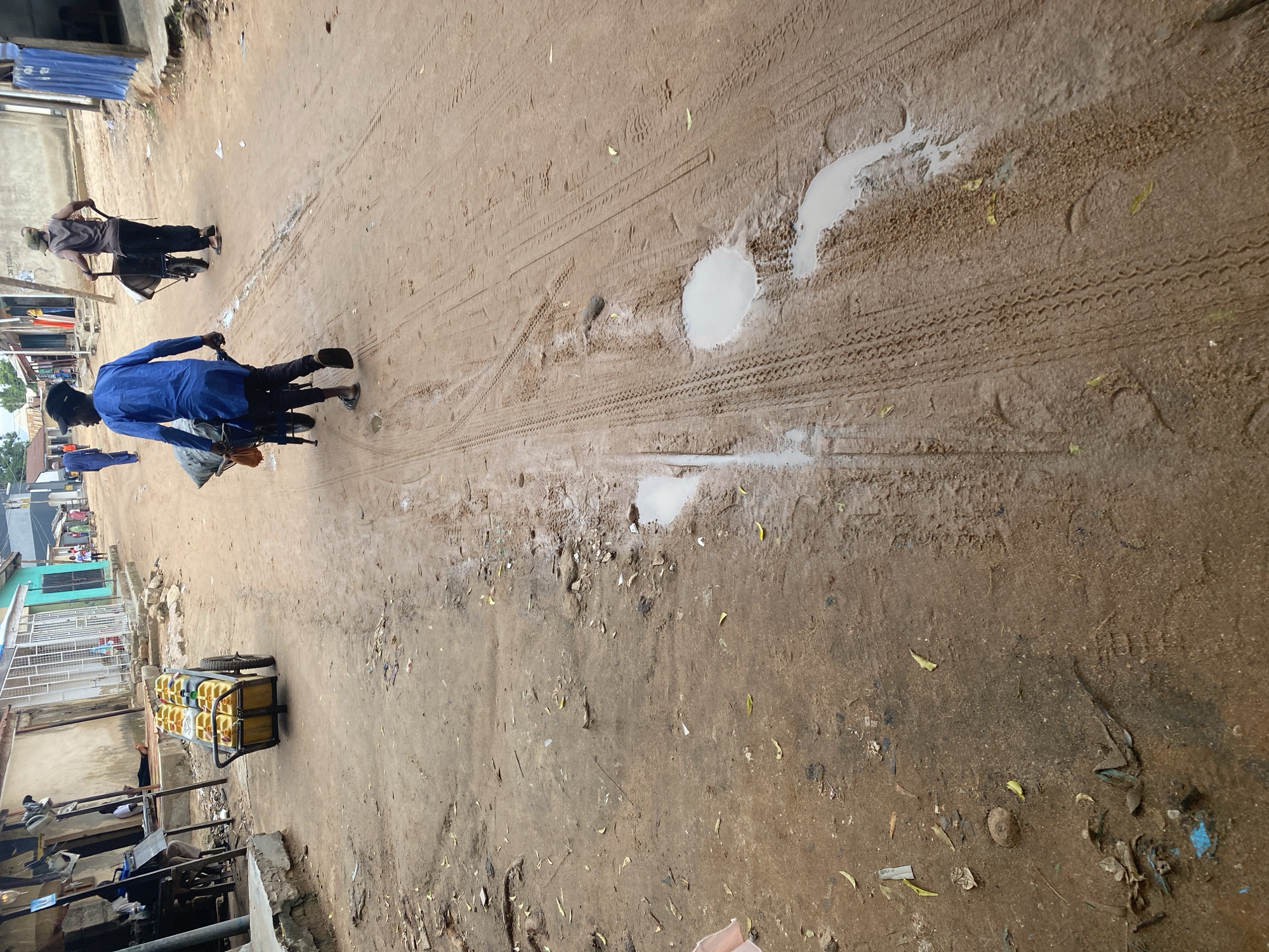 This media file shows an outdoor scene in Jalingo, Nigeria, on a dirt road with puddles and tire tracks. Two men are on a motorbike in the foreground, with the rider in a blue shirt and the passenger in a brown shirt. The motorbike is pulling a cart loaded with yellow jerry cans. Further down the road, another person is on a similar motorbike with a cart, but their load is not clearly visible. The surroundings suggest a residential or commercial area with buildings lining the road. The time of day appears to be daytime, and the weather seems overcast, as indicated by the puddles and the lack of harsh shadows. The overall atmosphere is one of daily life and activity in a Nigerian town. No visible text is present in the media file.