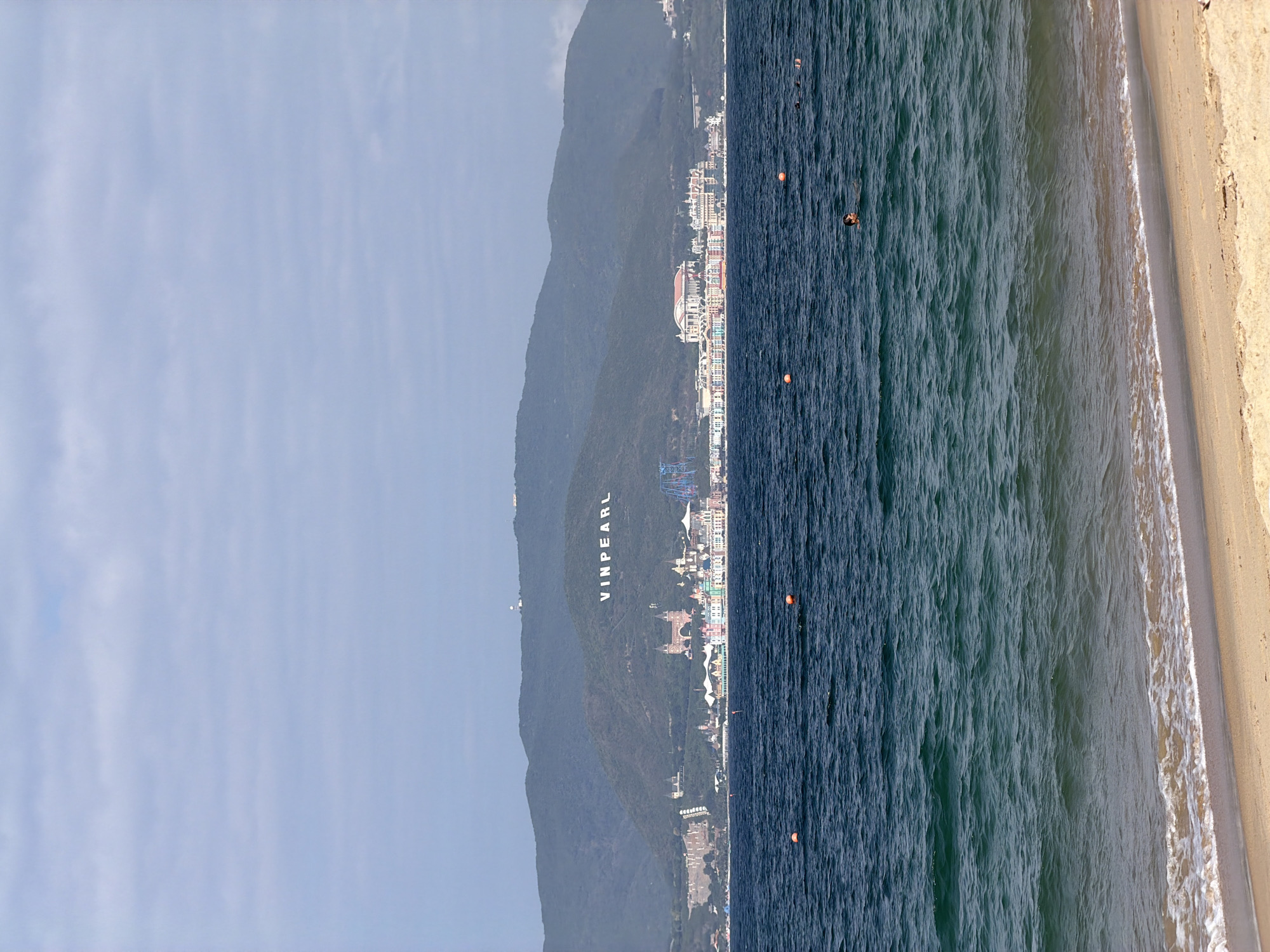 The image presents a wide outdoor view of a coastal scene, identified as being in or visible from Nha Trang Ward, Vietnam, showcasing the Vinpearl resort complex.
In the foreground, a strip of light brown sand beach occupies the bottom of the frame, where small white-crested waves are breaking. The immediate water transitions from a lighter green-blue near the shore to a darker blue-green further out, exhibiting visible surface ripples. Two orange spherical buoys are present in the water, positioned at different depths. At least four individuals are swimming or floating in the ocean; one person's head and shoulders are clearly visible in the mid-foreground, facing towards the left.
The midground features a large mountainous landmass, densely covered in green vegetation, rising from the sea. Prominently displayed on one of the mountain slopes are large white capital letters spelling "VINPEARL". Along the base of this mountain, stretching across the coastline, is an extensive resort and theme park complex. This complex comprises numerous multi-story buildings exhibiting varied architectural styles and colorful facades, including castle-like structures with pointed roofs, a grand building with a classical white facade and a red roof, and visible amusement park elements such as a tall blue roller coaster.
The background consists of an overcast sky, characterized by pale blue-grey, horizontally stratified clouds.