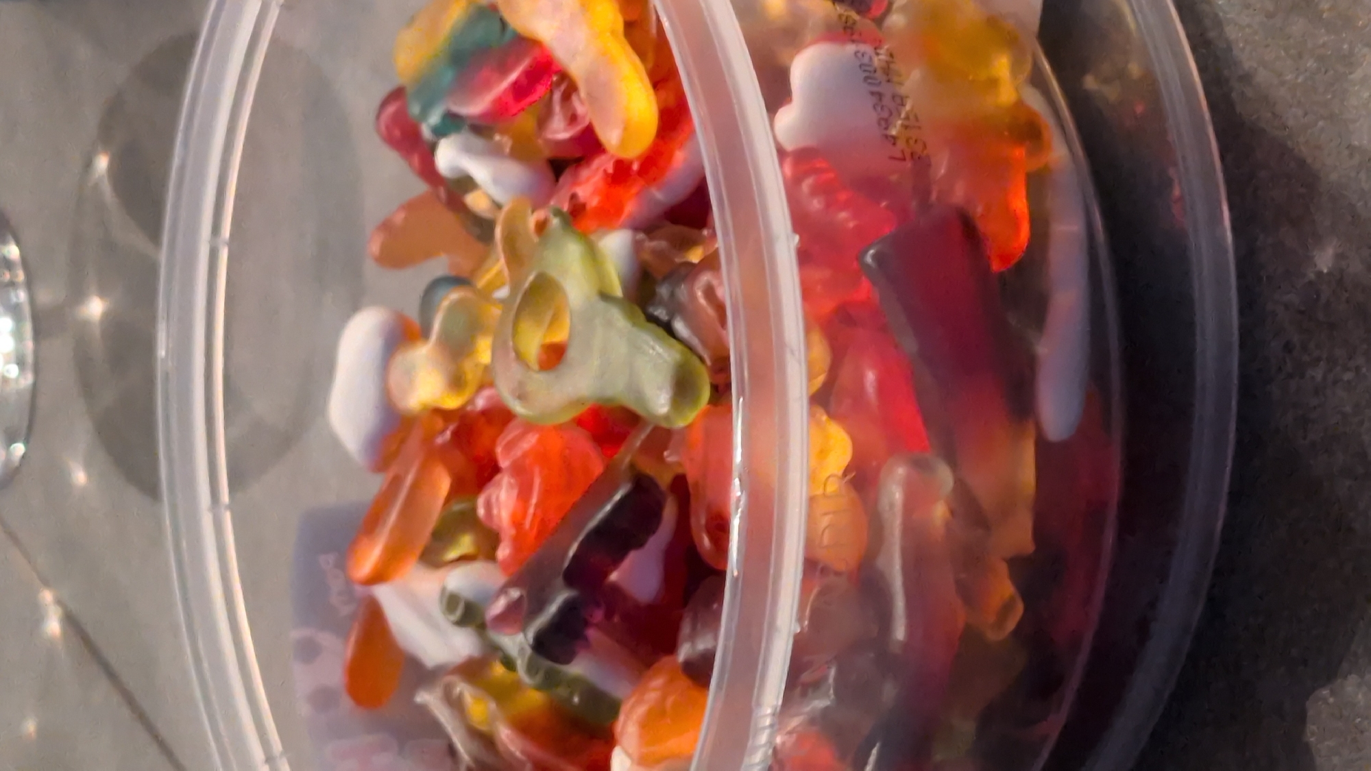 This is a close-up, slightly angled, overhead shot of a clear plastic container filled with various colorful gummy candies. The candies are assorted in shape and color, with some appearing as gummy worms, bears, and other novelty shapes like airplanes and keys. The lighting suggests it might be daytime, as evidenced by the bright reflections and shadows on the greyish surface beneath the container. The container itself has a ribbed rim for sealing.
Some of the candies are translucent and brightly colored in shades of red, orange, yellow, green, and blue. The texture of the candies appears soft and gelatinous. A faint, blurry label with some text is visible on one of the red candies in the upper right portion of the container, though the text is not clearly legible. The overall scene is a simple, everyday one, likely a snack or a treat being stored or displayed. There are no people visible in the frame. The setting appears to be indoors or outdoors on a flat surface.