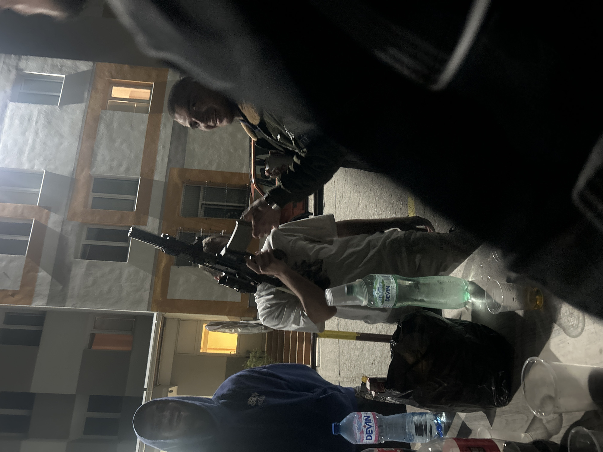 The image shows a group of people gathered outdoors at night. One individual holds what appears to be a firearm. Two people can be seen standing in the foreground, while another wears a blue hoodie at the bottom of the frame. There are drinks, bottles of water (Devin), and plastic cups on a table, and a black bag. The background includes a building with windows. I cannot identify the precise location in Veliko Tarnovo, Bulgaria.