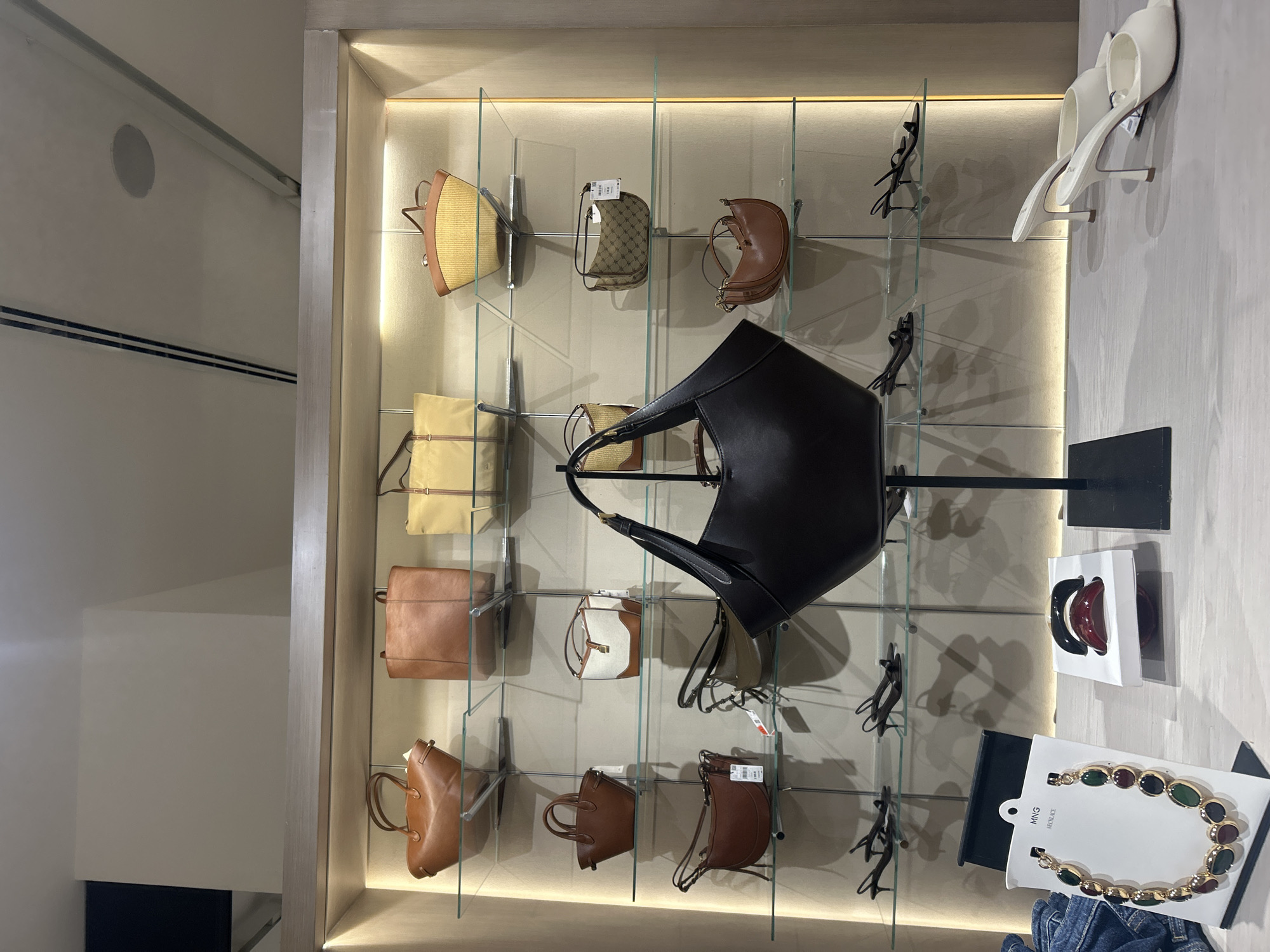 The image shows a display case in a retail setting, likely a store selling handbags and accessories. The location is unspecified, the city of San Bernardino Tlaxcalancingo, Mexico is not determined. The display case is filled with a variety of handbags in different styles, colors, and materials, arranged on glass shelves. Also visible are accessories such as shoes, hair accessories, and a necklace displayed alongside the handbags. The overall environment is well-lit and organized, suggesting a typical retail store interior. No people are visible in the image.