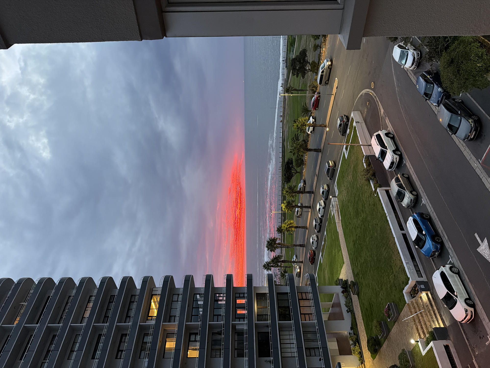 View from a high vantage point over the city of Cape Town, South Africa. The foreground shows the facade of a modern apartment building with repetitive balconies. Beyond the building, a coastal scene unfolds with a narrow strip of beach dividing the ocean from a green parkland with palm trees and a road. Cars are parked along the roadside. A band of red-orange sunset is reflecting off the ocean with a cloudy sky above. The time of day is early evening. No people are distinctly visible. The image is taken from inside a building looking outwards.