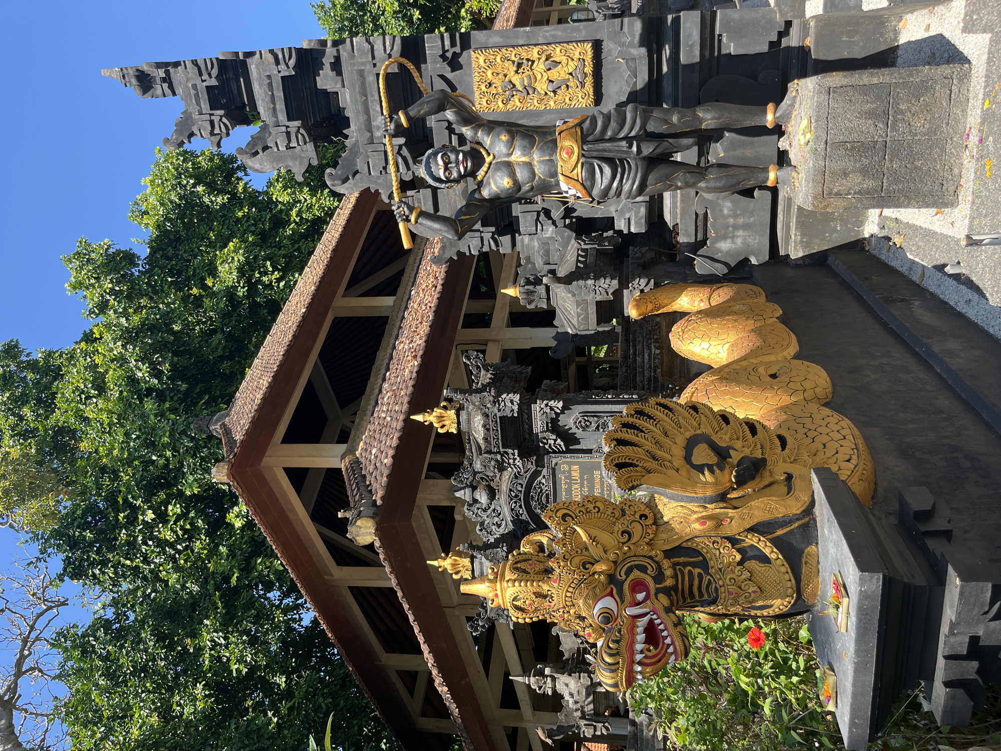In Kuta Selatan, Indonesia, a vibrant temple scene unfolds under a clear blue sky, framed by lush green trees. The foreground is dominated by an ornate, gold-and-black dragon statue with a fearsome expression, its body coiled and adorned with intricate details. Beside it, a dark-skinned statue of a muscular figure stands tall, holding a whip-like object above its head. This figure sports a red mouth, yellow jewelry, and a golden belt. Behind these statues, a traditional temple building with a brown, tiled roof is visible, its architecture featuring exposed beams and intricate carvings. Several smaller, dark statues with golden accents flank the entrance to the temple. The ground around the statues is covered in gravel and fallen leaves, with offerings of flowers and incense placed on small platters in front of the dragon sculpture.