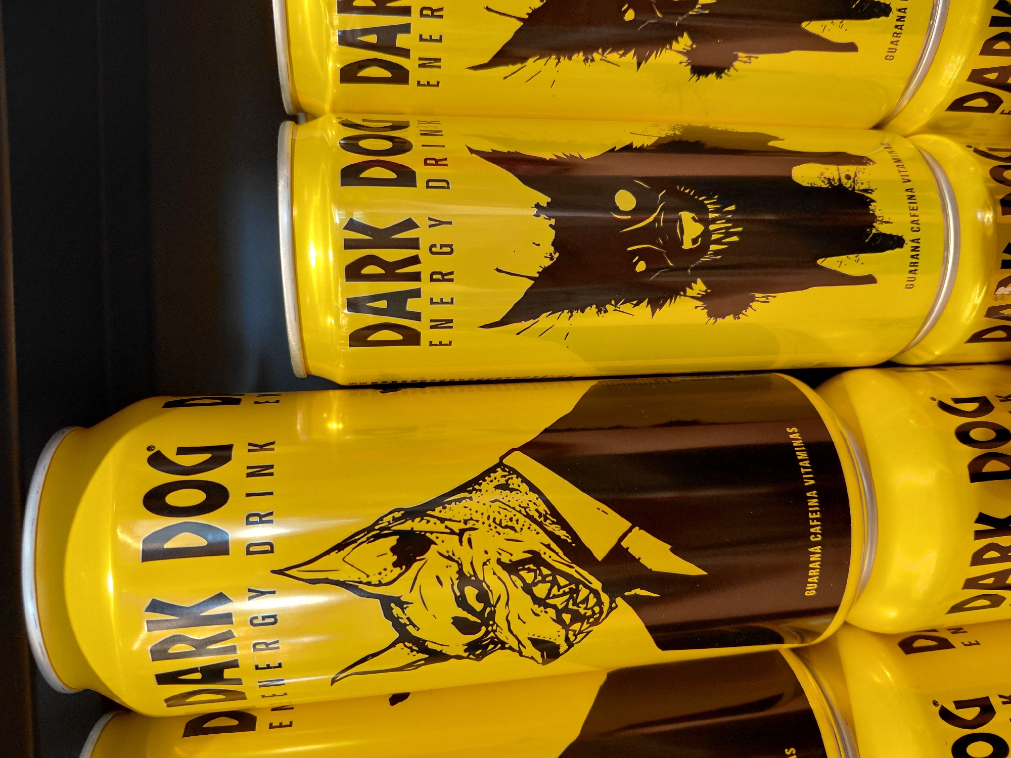 This is a close-up, vertical photograph of several yellow cans of "Dark Dog" energy drink stacked on a shelf. The cans are predominantly yellow with black graphics. The logo "DARK DOG" is prominently displayed in large black letters, followed by "ENERGY DRINK" in smaller text. Each can also features a black silhouette of a stylized dog's head with sharp teeth and spiky fur. Some cans also have the text "GUARANÁ CAFEINA VITAMINAS" which translates to "Guarana Caffeine Vitamins". The lighting suggests an indoor setting, possibly a store or supermarket, with the cans arranged neatly for display. The image appears to have been taken during the day due to the brightness and reflections on the cans. The overall impression is a product advertisement or a consumer photograph of a retail product.
