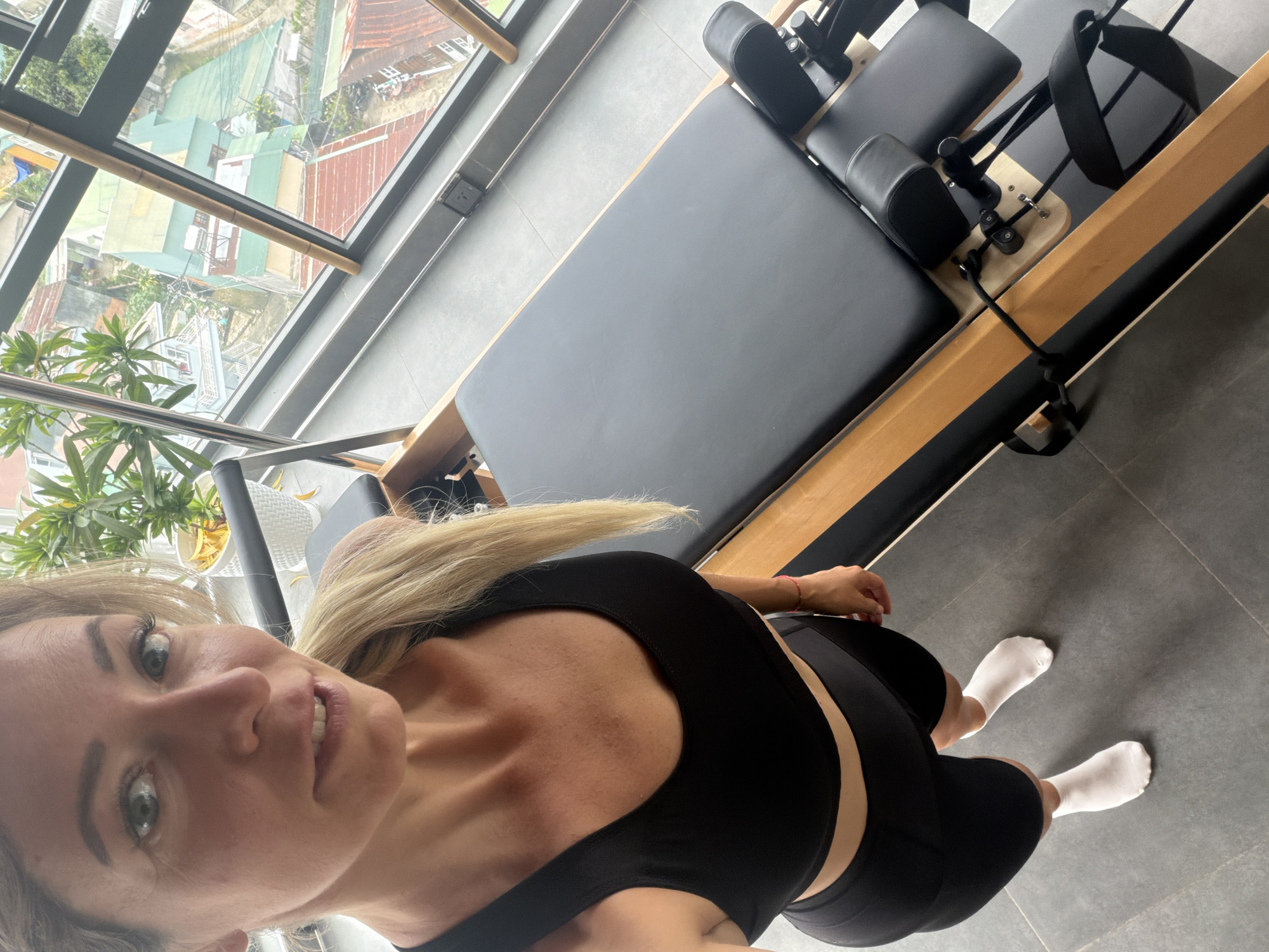 A woman with blonde hair pulled into a ponytail, wearing a black sports bra and black athletic shorts, with white socks, stands in a room with large windows overlooking a city. She is looking at the camera and appears to be taking a selfie. In the foreground, there is a Pilates reformer machine with a black carriage and wooden frame. The room has a gray tile floor and a plant in a white pot is visible near the window. The city view outside the window shows buildings with red and green roofs, suggesting an urban environment. The overall scene appears to be a home gym or a studio in Da Lat, Vietnam, given the architectural style of the buildings visible outside the window.