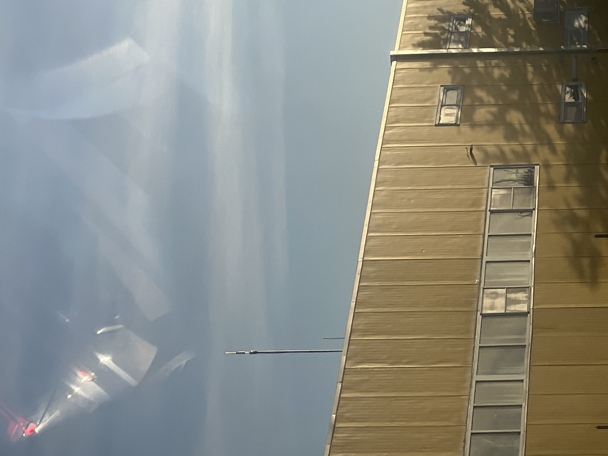 The image shows the side of a large, yellow-paneled building with a row of windows running horizontally across its facade. A tall, thin antenna stands against a clear blue sky, which is partially obscured by reflections and light streaks. Shadows of trees are cast across the building, indicating it is daytime and sunny. The building appears to be located in an industrial or commercial area, with no immediate signs of residential activity or prominent landmarks. The town of Dragushinovo, Bulgaria, is mentioned as the location, but no specific features within the image confirm this.