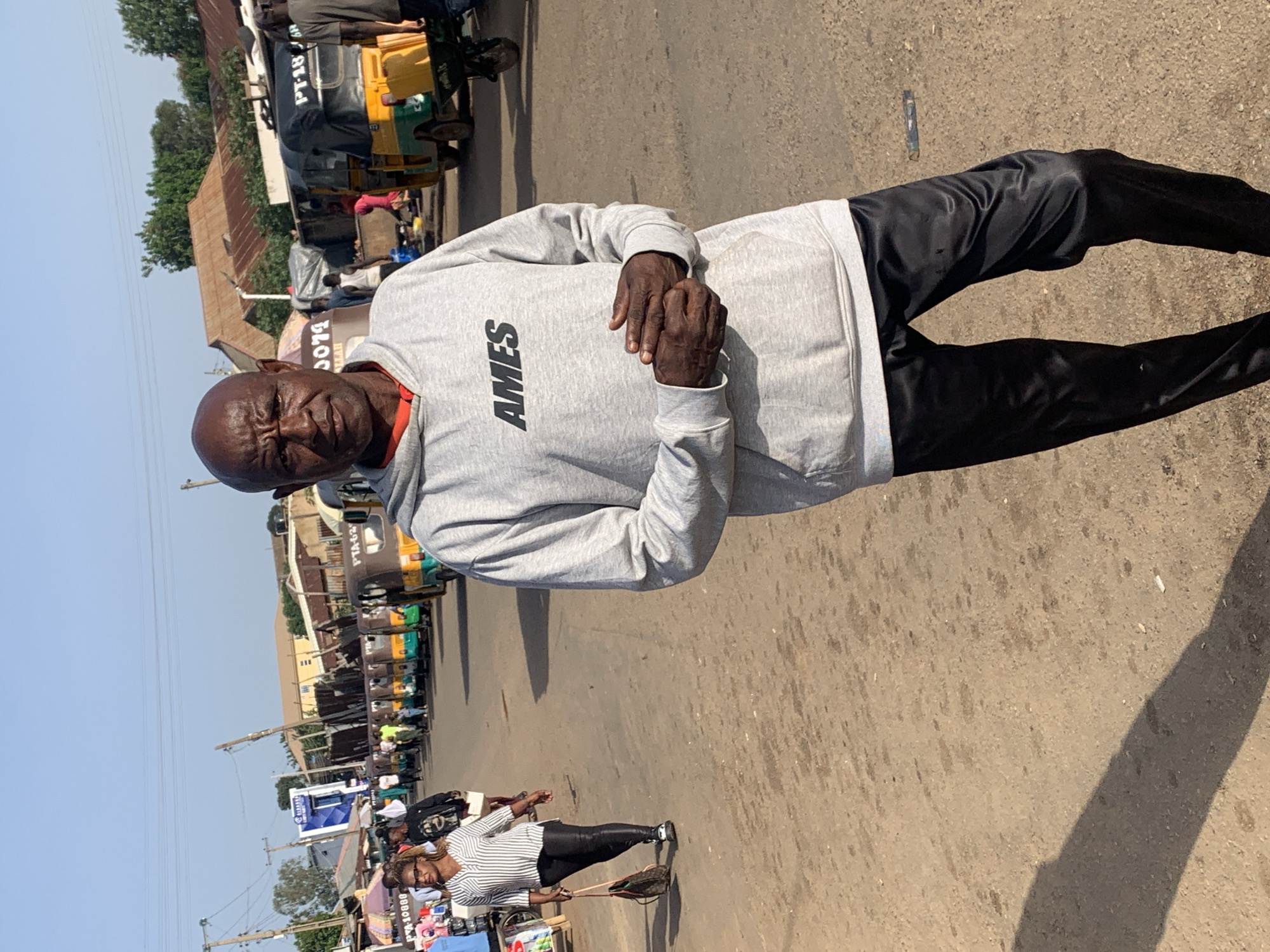 The image shows a portrait of an elderly Black man in the foreground, looking directly at the camera with a stern expression. He is wearing a light gray hooded sweatshirt with "AMES" printed in black lettering on the chest and black pants. His hands are clasped in front of him. The man is standing on a dusty, unpaved road.
In the background, a busy street scene unfolds. Several three-wheeled vehicles, commonly known as "keke napeps," are parked or moving along the road, some of them brightly colored yellow and green. There are also market stalls and buildings lining the street, with some corrugated metal roofs visible. People are walking and interacting in the background, suggesting a bustling marketplace or transportation hub. The overall impression is of a daytime scene in a warm climate, likely in Nigeria, given the context provided. The lighting suggests it is a sunny day. The scene depicts everyday life in a lively urban environment.
