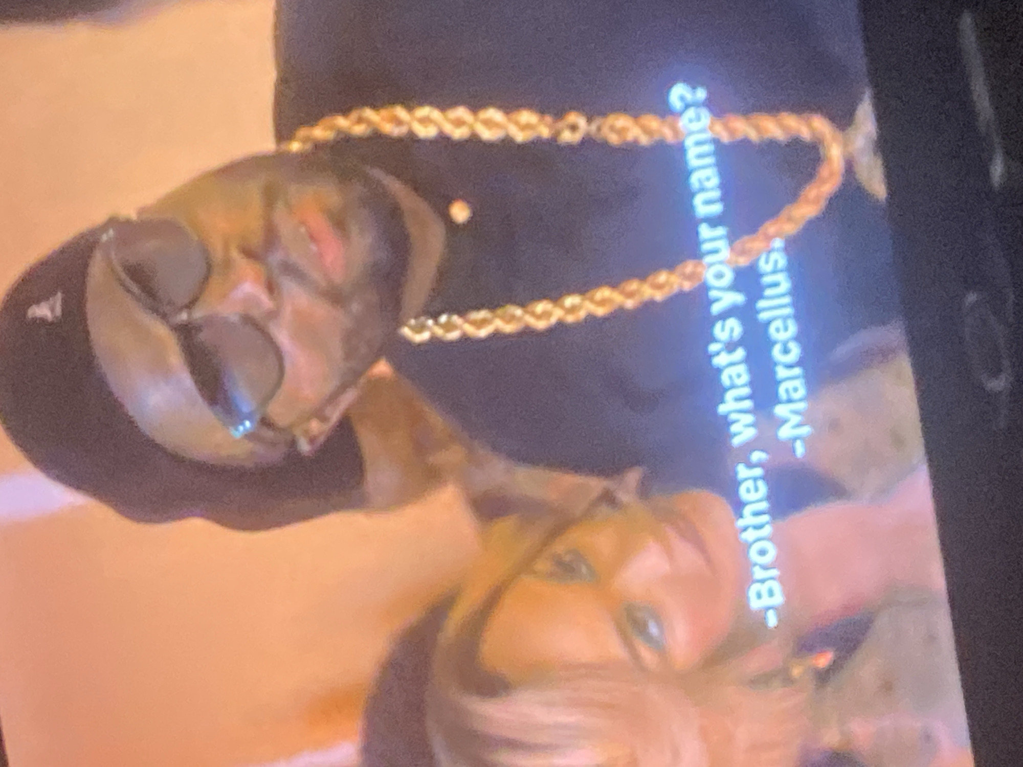 An image captures a rotated scene from a screen, displaying two individuals and subtitle text. The image is rotated approximately 90 degrees clockwise. In the upper part of the frame, a man with dark skin is visible, wearing dark sunglasses, a dark baseball cap, and a thick, gold-colored chain around his neck. He has facial hair. His gaze is directed downwards and to the left (relative to the original image orientation).
Below him, partially obscured, is a woman with blonde hair, looking upwards. Her features are less distinct due to blurriness, but she appears to be light-skinned. The background is softly blurred with warm, orange-brown tones, suggesting an indoor setting.
Subtitle text is prominently displayed on the screen, indicating a dialogue:
"-Brother, what's your name?"
"-Marcellus."
The image appears to be a photograph of a digital display, evidenced by the visible text and the overall quality, suggesting it's a still from a movie, TV show, or video.