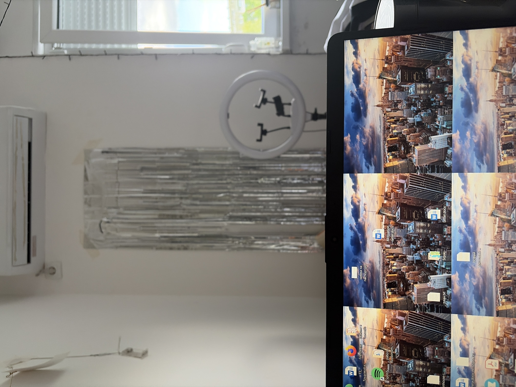 The image displays an indoor scene, likely a room within a residence in Vozdovac Urban Municipality, Serbia. No people are visible.
The room features white walls. At the top, a white-framed window with horizontal blinds allows daylight to enter. Below the window, a string of small, decorative lights is draped. To the left, a white air conditioning unit is mounted on the wall, with an electrical outlet visible beneath it.
Dominating the center of the wall is a shiny, silver, tinsel-like decoration or curtain. Above this, a white ring light with two phone mounts attached is visible. In the lower-left section of the wall, a white, folded object resembling a paper airplane is attached, with a thin wire extending from it to a small white device.
On the right side of the image, a tablet or monitor is shown in portrait orientation, displaying a desktop interface. The background image on the screen is a city skyline under a dramatic sky with clouds, suggesting either dusk or dawn. Several desktop icons and files are visible, including Microsoft Word documents, applications, and folders.
Visible text from the screen includes various file names and application labels. These include "заявка на паспорт.jрд" (application for passport.jpg), "Motivation latter.docx" (likely "Motivation letter.docx"), "план.txt" (plan.txt), "Srpski" (Serbian), "Letter.docx", "Новый текстовый" (New text file), "Firefox.exe", "резюме и письмо" (resume and letter), "работа Roobinium" (work Roobinium), and "Spotify". The presence of the word "Srpski" further corroborates the location in Serbia.
