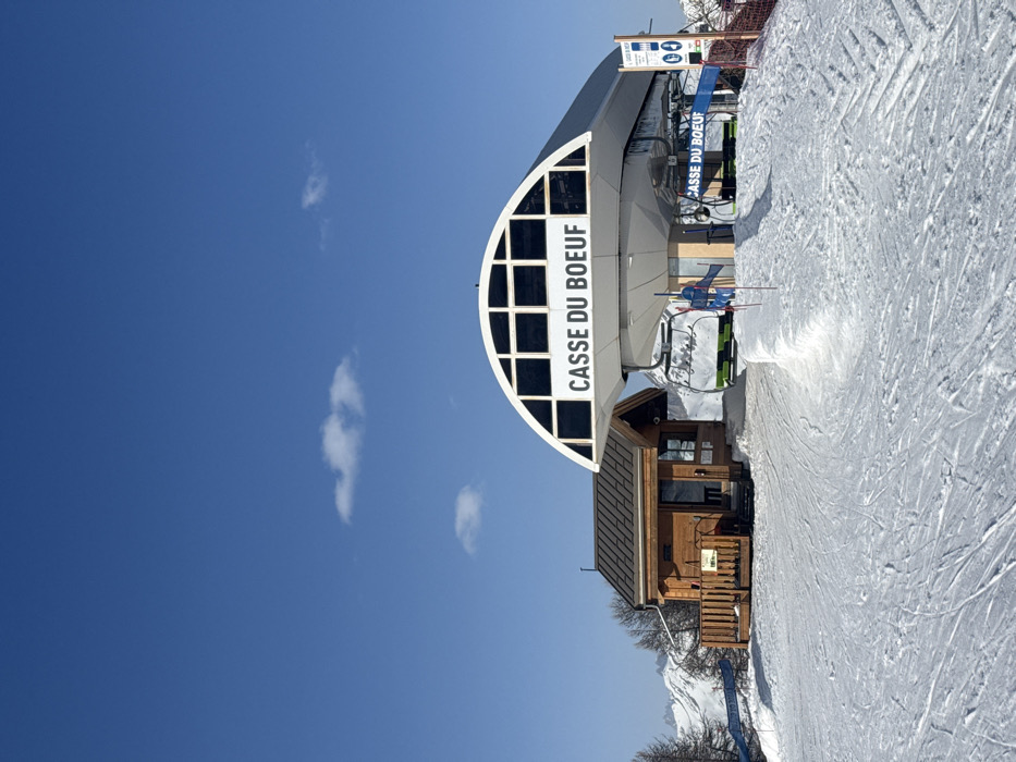 The image displays the "Casse du Boeuf" ski lift station located in a snow-covered mountain environment within the ski resort area of La Salle-les-Alpes, France. The central subject is a large, modern ski lift terminal featuring a prominent white, arched roof structure with the name "CASSE DU BOEUF" displayed in dark capital letters. Inside the station, multiple ski lift chairs, distinguished by green and black seats, are visible. The loading area in front of the station is demarcated by blue barriers and red flags. To the left and slightly behind the main lift station, a smaller, rustic wooden building with a dark, pitched roof and an attached wooden deck is present. The foreground and midground are covered in groomed snow, showing visible ski tracks. In the background, under a clear blue sky with sparse white clouds, snow-capped mountains and scattered leafless trees are visible. No people are observed in the scene.
