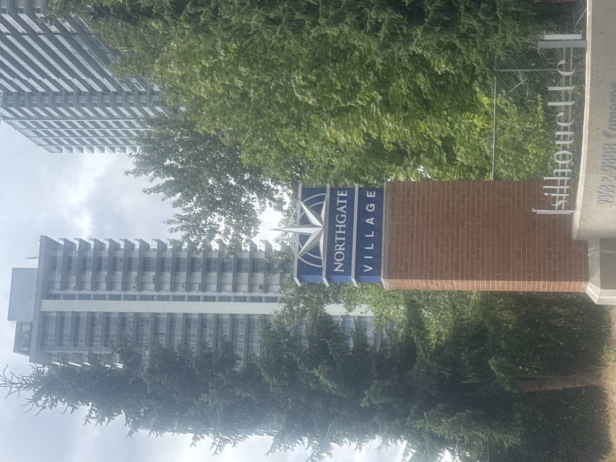 A vertical, eye-level outdoor shot captures a brick monument with a blue sign reading "NORTHGATE VILLAGE" topped with a white starburst emblem, located in Burnaby, Canada. Behind the monument, two tall residential buildings rise against a cloudy sky. Lush green trees, including a tall evergreen on the left, partially obscure the view of the buildings. A metal fence is visible behind the monument and trees. At the base of the monument, a silver cursive sign reads "silhouette" above a concrete base with engraved numbers "9868 & 9888 Cameron." The scene is bathed in overcast daylight, creating a muted color palette.