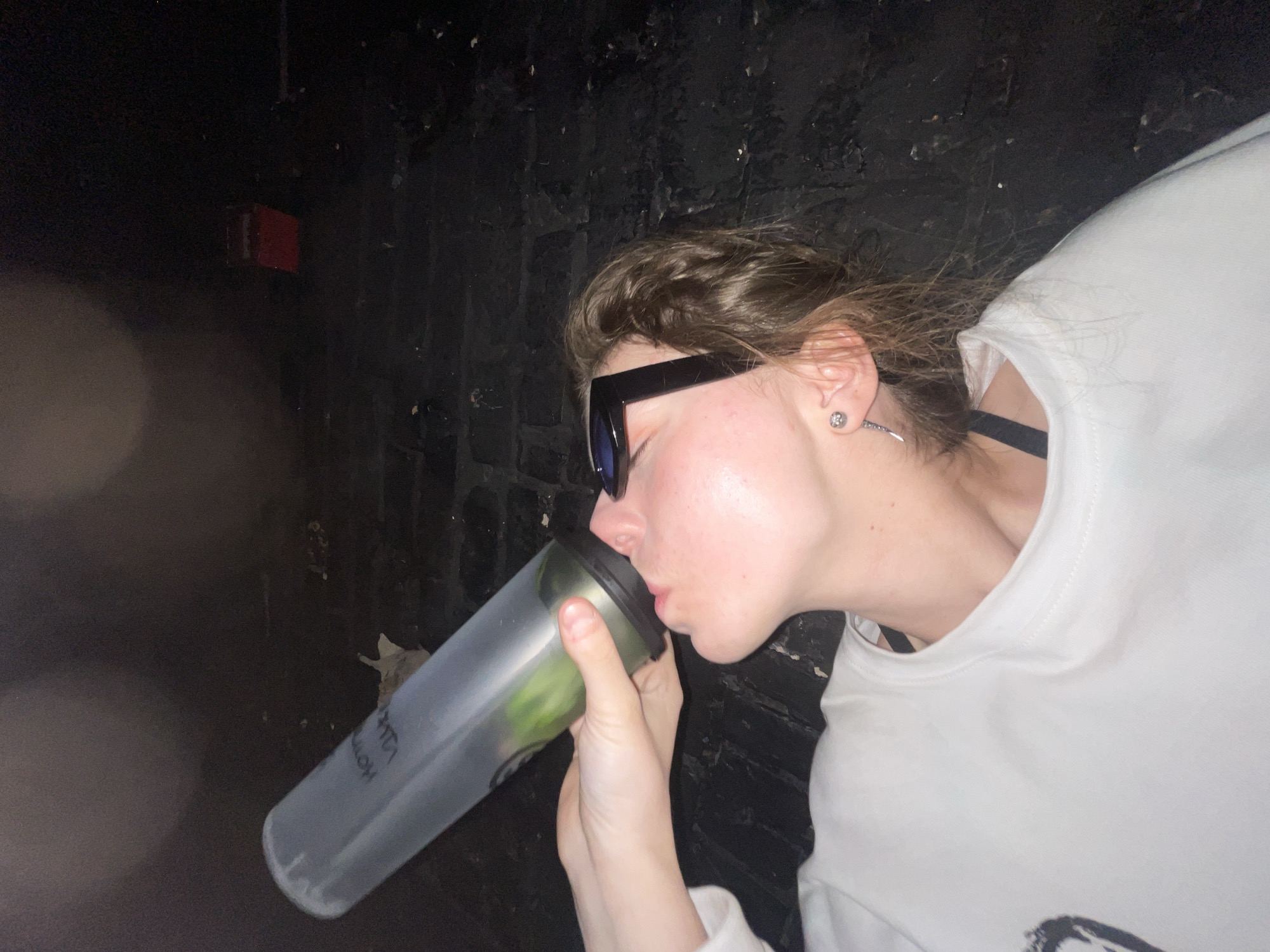 The image displays a close-up, horizontal view of a person drinking from a large clear bottle. The subject, a young adult, is seen in profile, with their lips pressed against the bottle's spout. They wear dark sunglasses that obscure their eyes, and their medium-brown hair is slightly disheveled.
The person is dressed in a white long-sleeved top or sweatshirt, with a black bra strap visible on their shoulder. They accessorize with two silver earrings: a stud on one earlobe and a longer, pointed design on the other. Both hands are holding the clear bottle, which appears to contain slices of green fruit or vegetable, like cucumber or lime, in water. Faint text is visible on the bottle's side, partially legible as "HOM" and "LOW."
The background consists of a dark, textured wall, suggesting an indoor setting with dim lighting, possibly a club or bar. A small, red rectangular object, resembling an alarm or electrical box, is visible in the upper left background. The overall mood is casual and focused on the act of drinking. This scene is set in Moscow, Russia.