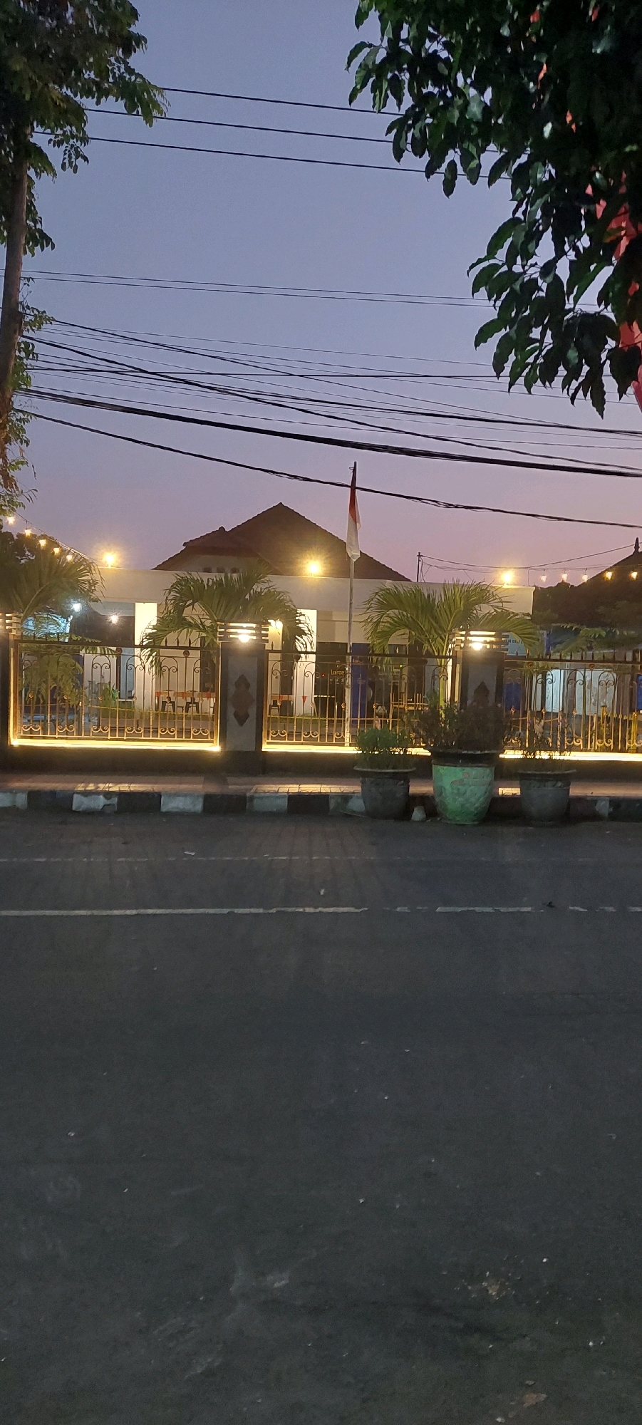 The image captures a street view in Lumajang, Indonesia, at dusk. In the foreground, a dark asphalt road with a white line is visible, bordered by a sidewalk with black and white markings. Behind the sidewalk, a decorative metal fence, illuminated by warm yellow lights, encloses a building complex. The building features white walls and a brown tiled roof, with several palm trees planted in front of it. A flagpole with the Indonesian flag is positioned in front of the building. In the background, a hazy sky transitions from light blue to purple, with power lines crisscrossing the frame. The scene appears to be peaceful and well-maintained, with no people or vehicles directly in focus.