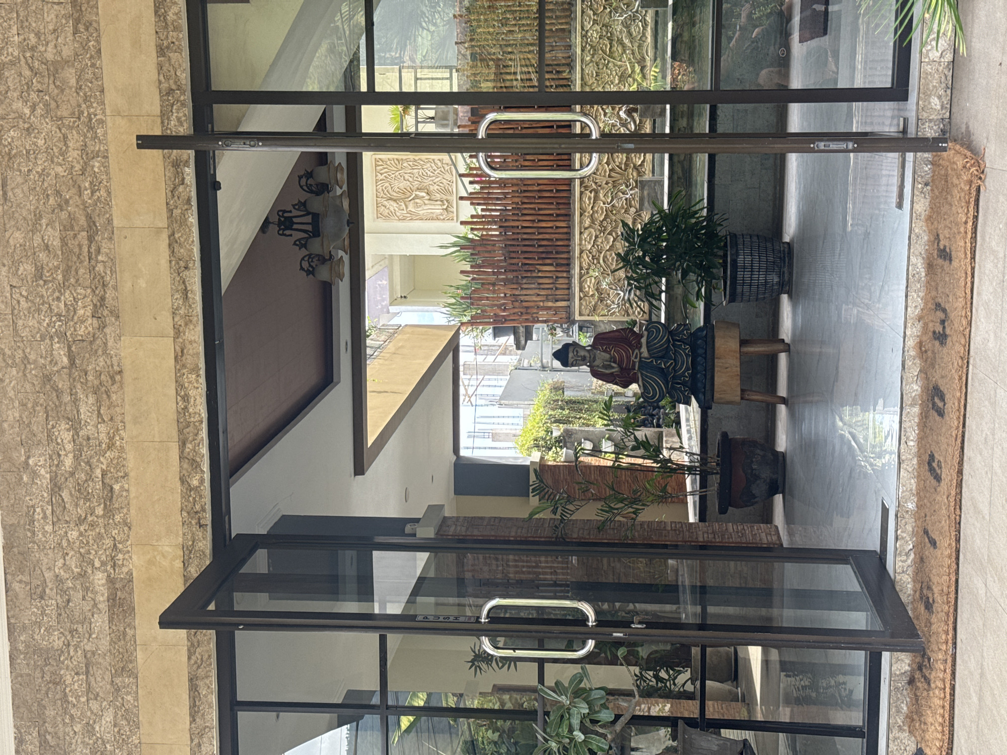 The image shows an open glass double door entrance to a building. A Buddha statue is positioned centrally within the doorway, seated on a wooden stool. The area behind the statue features a decorative wall with carved motifs and a bamboo fence. To the left and right of the Buddha are potted plants. The floor inside the entrance appears to be polished tile, reflecting the light. A coir doormat is visible on the exterior tiled ground, with partial lettering that seems to read "WELCO." The building's exterior walls are clad in textured stone and lighter-colored stone bands. The overall impression is one of a serene and decorative entrance. The location is identified as Abang, Indonesia.