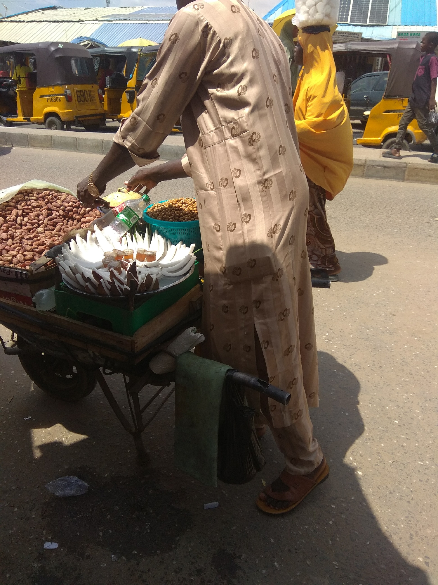 The image depicts a bustling street scene in Jalingo, Nigeria, under what appears to be a bright, sunny day. The primary subject is a person, likely a vendor, wearing a patterned beige robe and brown sandals. They are standing behind a cart laden with goods.
On the cart, several items are visible. A large bag of peanuts sits on the left, with a blue bucket filled with what appear to be chickpeas or similar legumes to its right. In the center, a platter is filled with artfully arranged pieces of white coconut, with small containers of what looks like honey or a similar sweet syrup placed among them. The cart itself is made of wood and metal, with a black wheel visible at the bottom.
In the background, several yellow auto-rickshaws, commonly known as "Keke napep," are parked or moving. One of these vehicles has the license plate "690K". A woman in a bright yellow hijab, with a white head covering, is also present, carrying a large bundle on her head. Further in the background, a young person walks by, carrying a plastic bag. Buildings with corrugated metal roofs and solar panels are visible, suggesting a commercial or residential area.
The overall impression is one of daily life and commerce in a vibrant Nigerian market. The sunlight casts distinct shadows, indicating it is likely daytime, possibly late morning or afternoon. The scene is lively and active, with the presence of multiple people and vehicles. There is no discernible text on the goods displayed, apart from the license plate on the Keke napep.