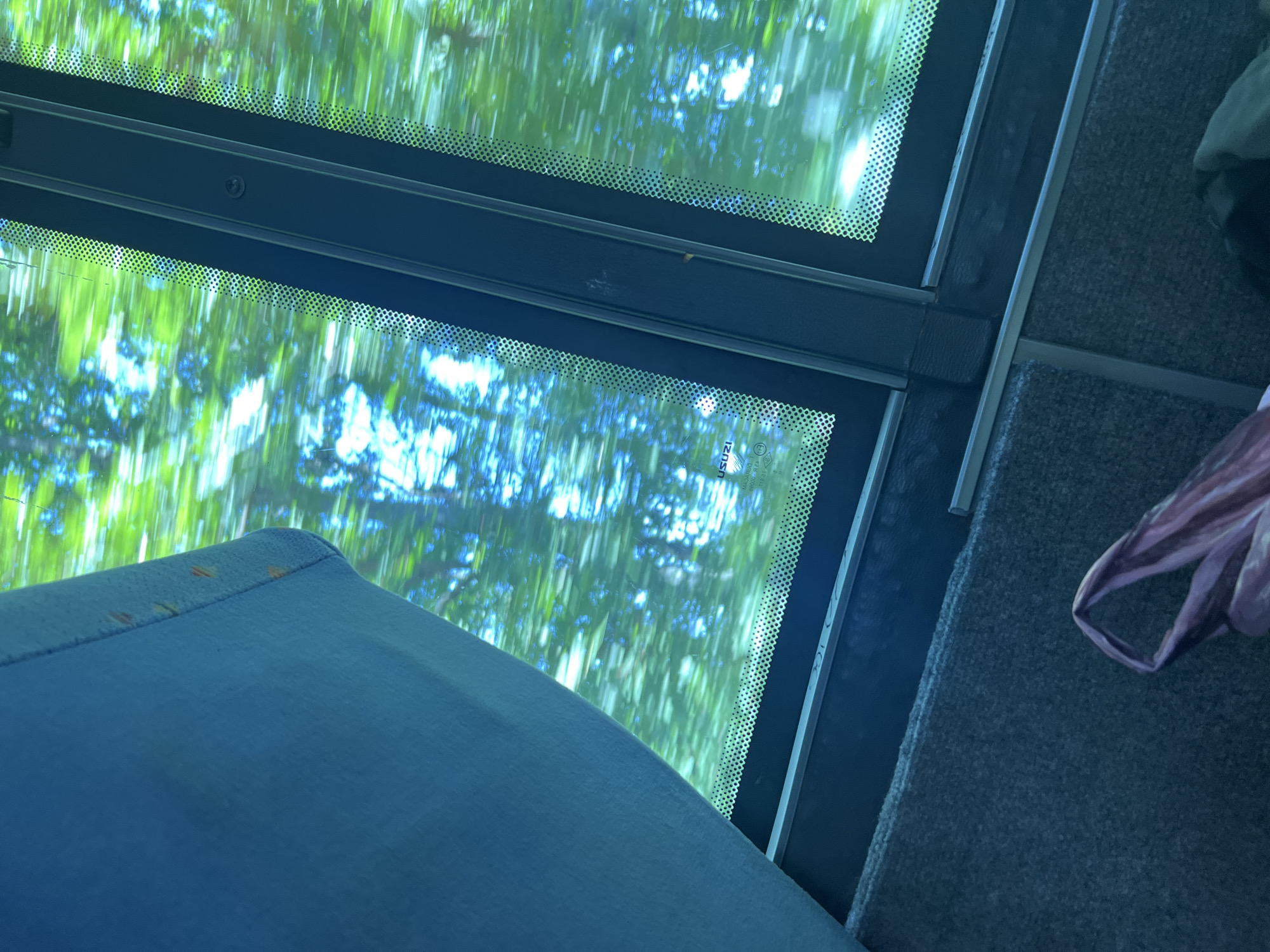 An interior view from a moving vehicle reveals a window offering a blurred perspective of green foliage and blue sky, indicating movement through a vegetated outdoor environment in Slivovo, Bulgaria. The upper-left quadrant is occupied by a blue, textured fabric, possibly a curtain or seat upholstery, exhibiting several small, faint, reddish-brown stains along its right edge. The central and right portions feature a multi-pane window framed by dark, structured elements. A vertical dark divider bisects the window, and a horizontal dark frame runs along its bottom. The window glass features a pattern of small dots (frit band) along its edges and displays partial text including "USUZI" and "E3 (43) R-0002", indicative of a manufacturer and certification. The bottom section of the image shows a grey, textured surface, likely carpet or floor upholstery, with a light-colored horizontal strip. A crumpled, translucent, light purple plastic bag is partially visible in the bottom right, next to an obscured dark, textured object. No individuals are depicted in the scene.