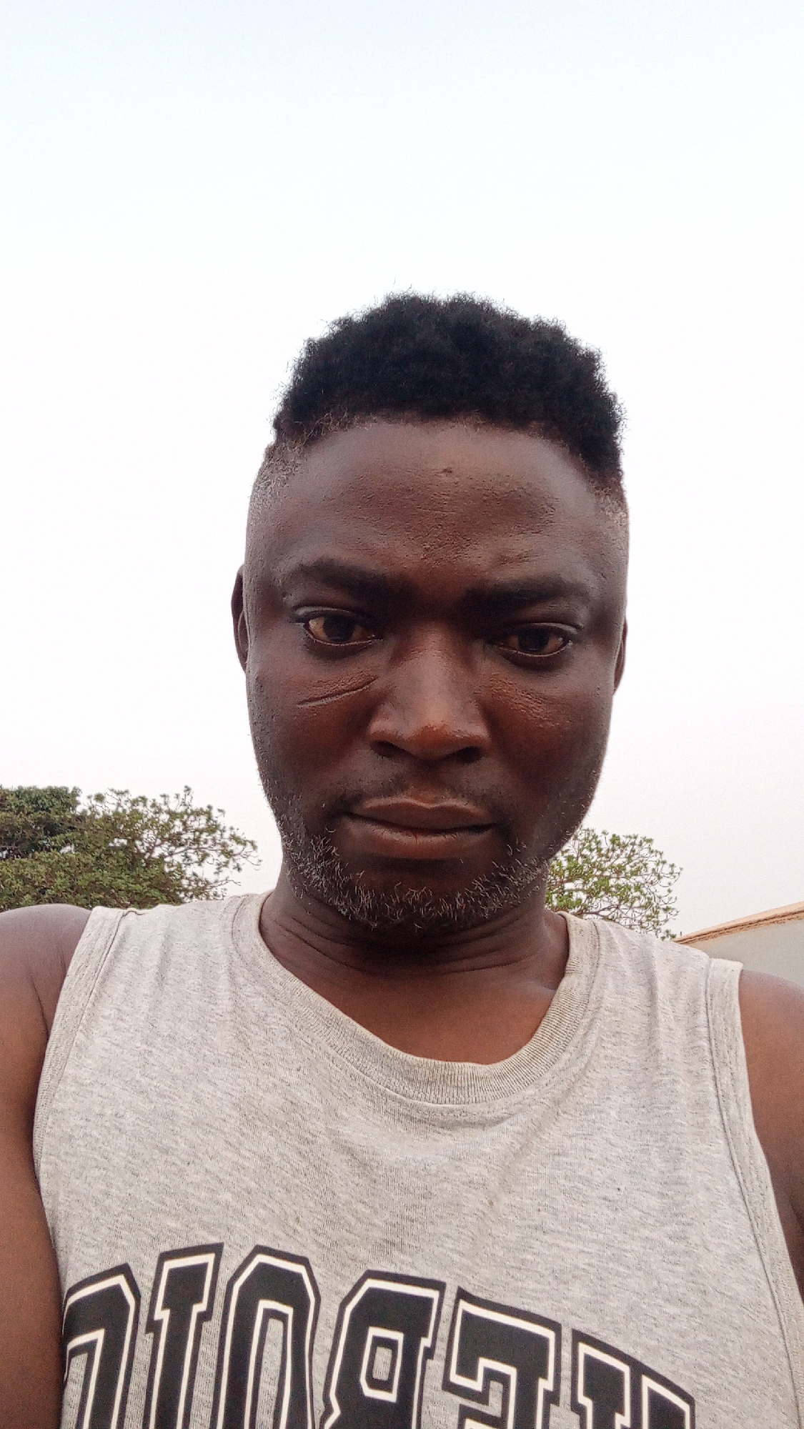 A medium shot captures a Black man, likely in his 30s or 40s, wearing a gray tank top with black and white lettering. He has short, dark hair with a faded cut, a beard with graying hairs, and a faint scar near his left eye. The background shows a cloudy or overcast sky, with foliage and what appears to be the corner of a building visible. This is presumably taken outdoors in the city of Television, Nigeria. The man looks directly at the camera with a serious expression.