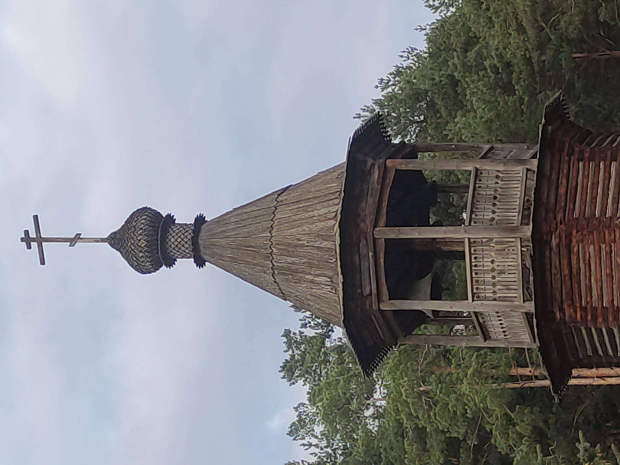 The image depicts the upper portion of a wooden Russian Orthodox church or bell tower, likely in a rural or forested area. The structure has a distinctive onion-shaped dome topped with a cross, a common feature of Russian Orthodox architecture. The dome and roof are covered in wooden shingles. Beneath the dome is an open-air bell tower with decorative wooden balustrades. The surrounding environment includes coniferous trees, partially obscuring the lower portions of the structure, and an overcast sky. The precise location is not discernible from the image.