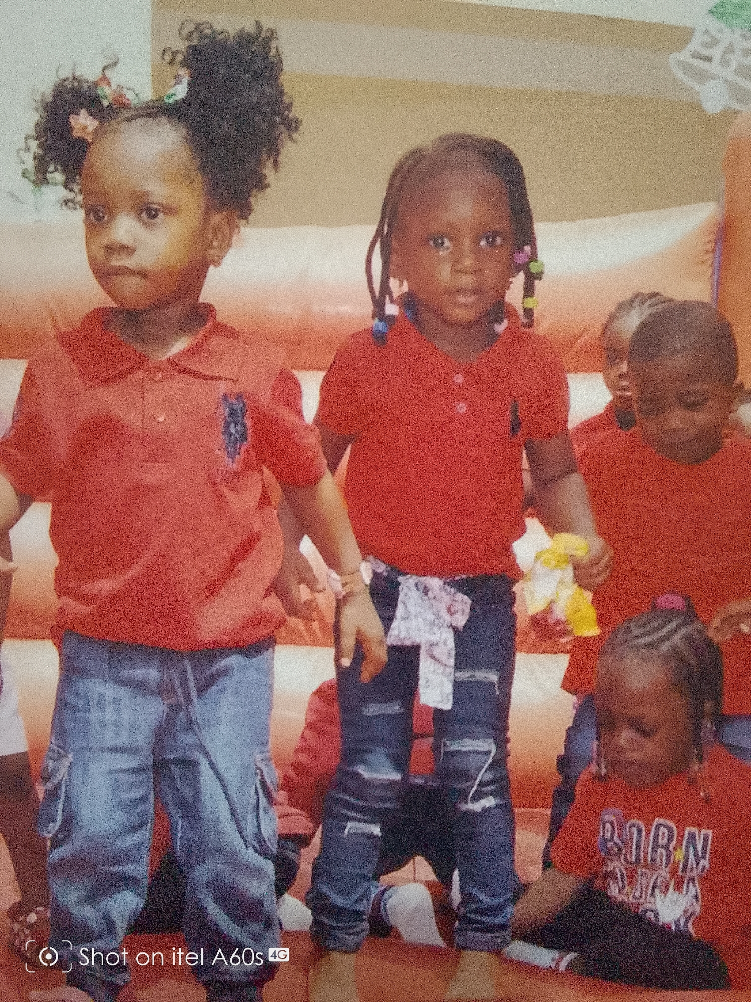 The image shows a group of children, presumably in Idimu, Nigeria, all wearing red polo shirts and blue jeans. The children appear to be on a bouncy castle or similar inflatable structure, indicated by the orange color of the background. The child on the left has two afro puffs tied with light-colored flowers. The child to her right has braided hair adorned with colorful beads. A white and gray patterned fabric is tied around her waist. At least three more children are visible in the background. A watermark "Shot on itel A60s 4G" is present at the bottom left corner of the image.