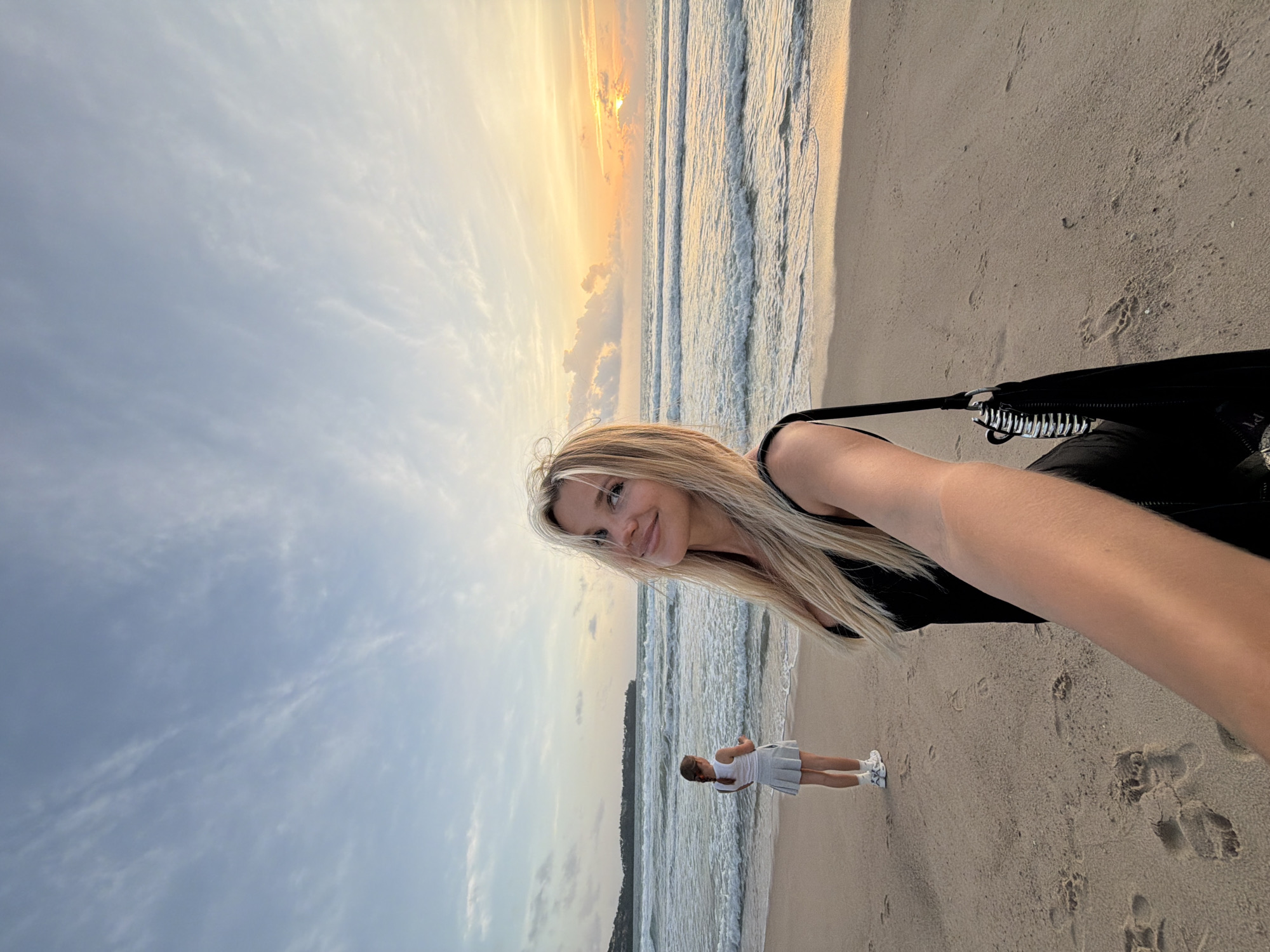 In this selfie, a woman with blonde hair smiles at the camera on a sandy beach. She is wearing a black outfit. To her left, a young girl in a white dress and sneakers stands facing the ocean. The ocean has gentle waves, and the sky is filled with clouds. The sun is setting, casting a warm glow over the scene, suggesting it is late afternoon or early evening. The beach appears to be a tropical location, given the mention of Choeng Thale, Thailand, which is known for its beautiful beaches. The overall mood is relaxed and happy, with the two individuals enjoying the seaside at sunset. There is no visible text in the image.