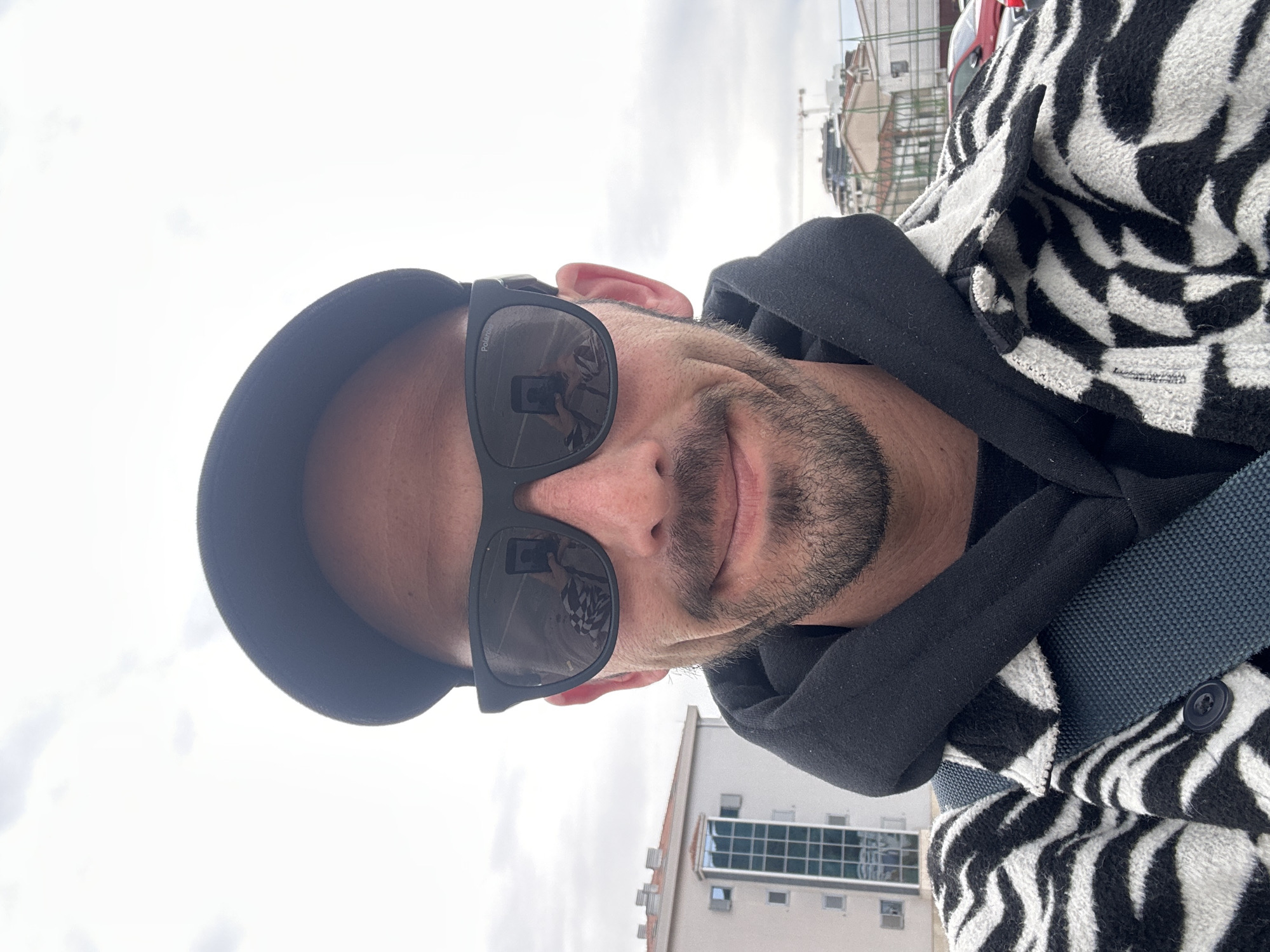 A man is taking a selfie outdoors. He is wearing a black cap, sunglasses with a reflection of the photographer holding a phone, a black hoodie, and a black and white zebra-striped jacket with a blue strap. He has a short beard and is smiling slightly. In the background, there are buildings, including a light-colored building with windows, and a cloudy sky. Based on the reflection in the glasses and the general style, the location is possibly Budva, Montenegro.