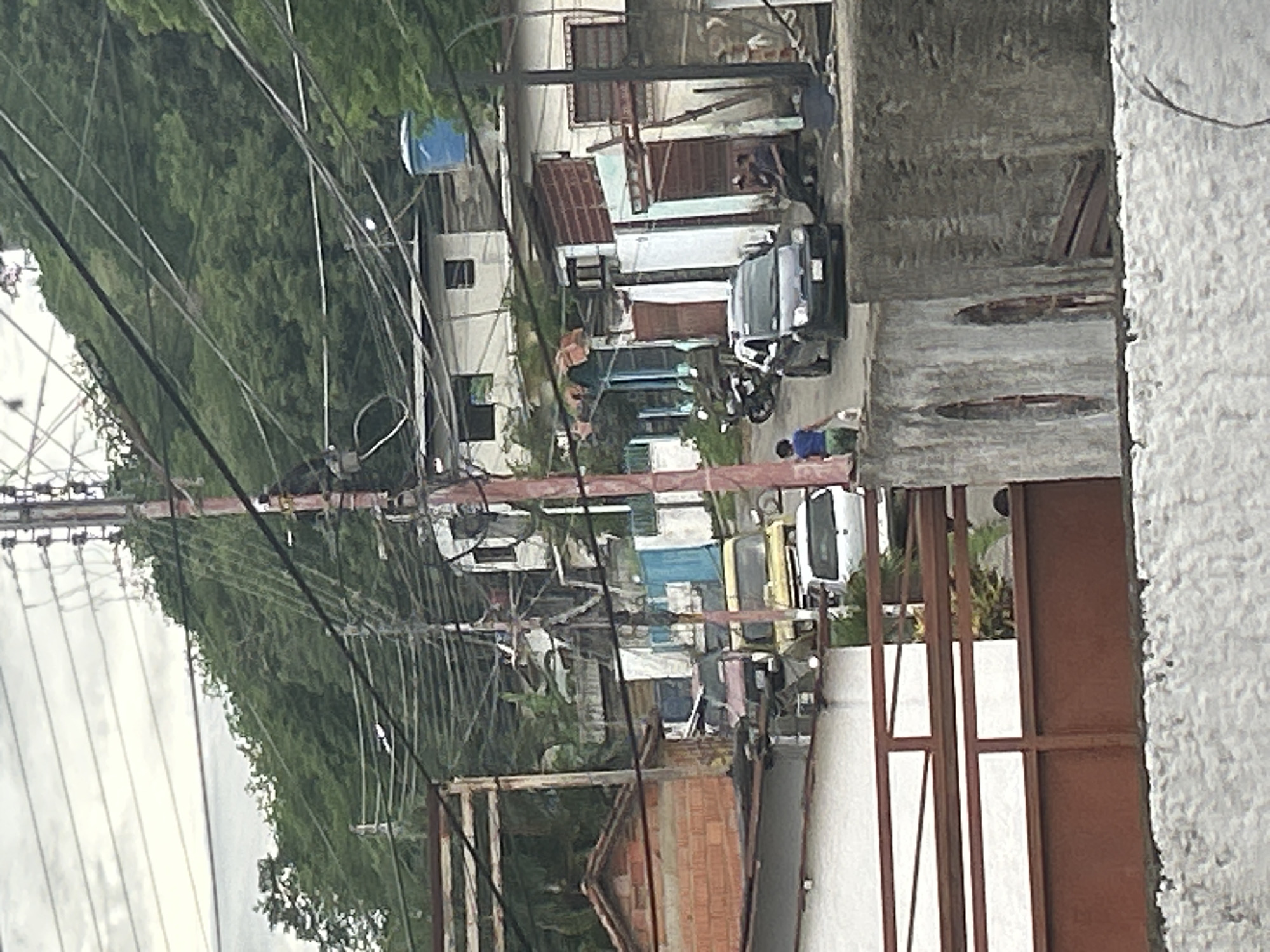 The scene shows a street in San Juan de Los Morros, Venezuela, taken from an elevated position, possibly a window or rooftop. The image includes several buildings, varying in construction and color. The buildings appear to be closely packed together. There are several vehicles parked along the street, including cars and a motorcycle. Power lines are strung across the scene. A densely forested area is visible in the background. People can be seen on the street, some appearing to be engaged in activities near the vehicles. The foreground features a brick structure, a metal gate, and a textured white wall, obscuring some of the street view.