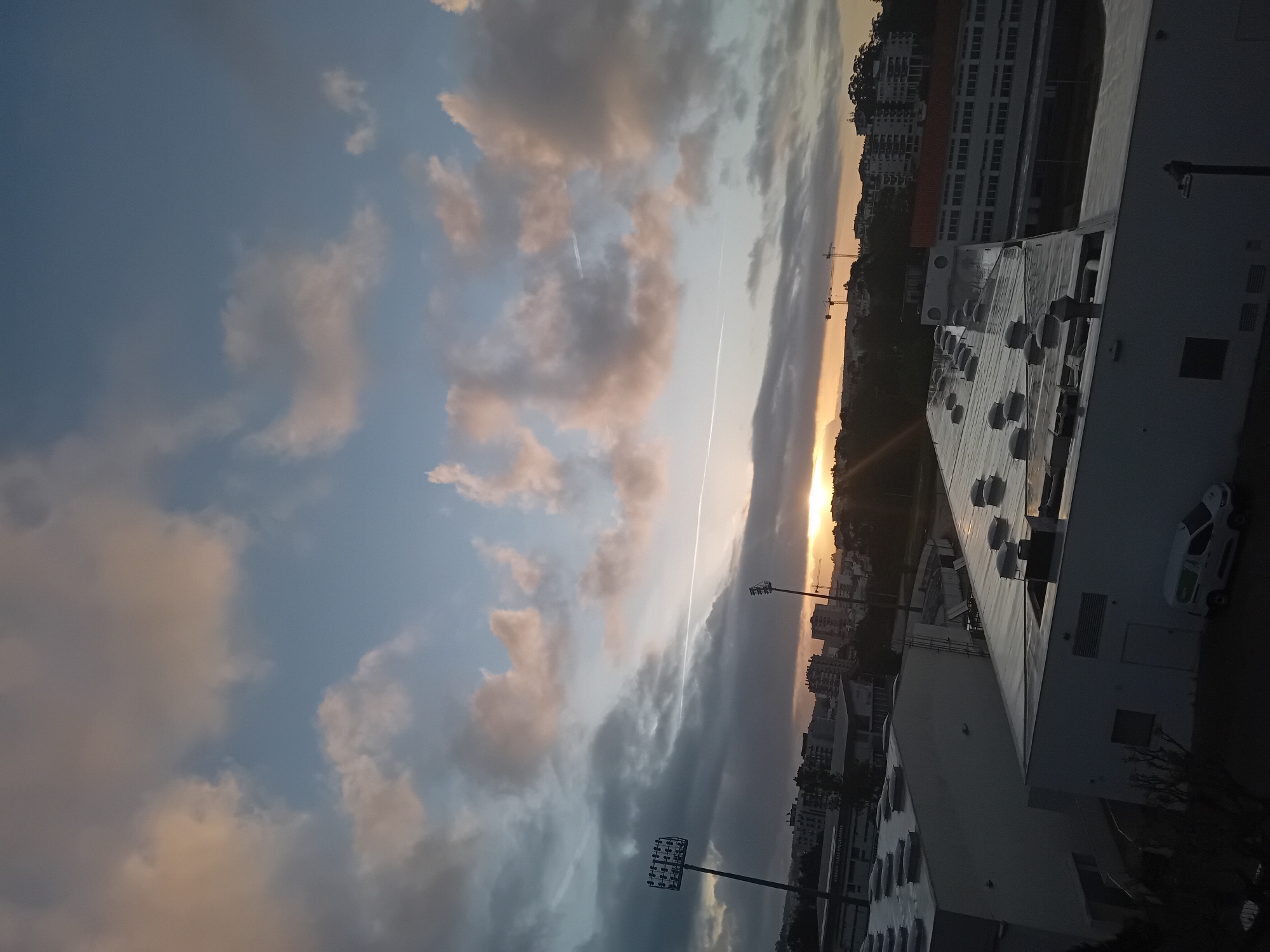 This media file, oriented horizontally, depicts an urban or suburban landscape at either sunrise or sunset. The sky occupies the majority of the frame, characterized by a dynamic interplay of blue and grey clouds, some tinged with warm orange and pink hues from the low sun. A distinct white contrail from an aircraft streaks diagonally across the sky, passing near the bright, partially obscured sun on the right horizon, which also exhibits a lens flare. A fainter contrail is visible further left.
In the lower portion of the image, several buildings are visible, often silhouetted or in shadow due to the backlighting. A large, flat-roofed building in the foreground on the right features numerous dome-shaped ventilation units or skylights on its roof. A white car, appearing to be a van or hatchback, is parked alongside this building. Further back, another building with many windows and what appears to be "PARKING PUBLICS" as indistinct text on its facade is present. Two tall floodlight poles, typical of sports venues, stand prominent, one closer and another further in the distance. In the background, numerous city buildings stretch across a hillside, and two construction cranes are visible near the horizon. The scene evokes a tranquil yet active urban environment transitioning between day and night.