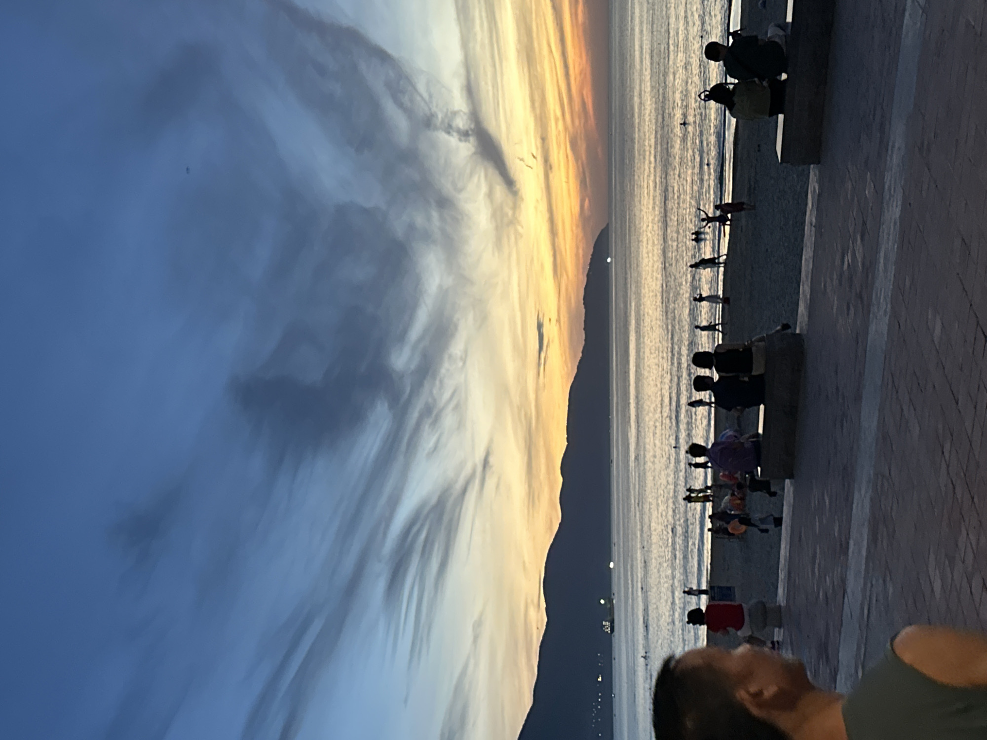 The image depicts a sunset scene at a beach in Da Nang, Vietnam. The sky is a gradient of blues and oranges, with wispy clouds. In the distance, mountains form a silhouette against the horizon, and a ship is visible on the water. A beach with people enjoying the evening is in the middle ground, with some individuals swimming or playing in the water, and others sitting on the sand or walking along the shore. In the foreground, a person is partially visible, looking out towards the ocean. The overall atmosphere is serene and peaceful, capturing the beauty of a coastal sunset.