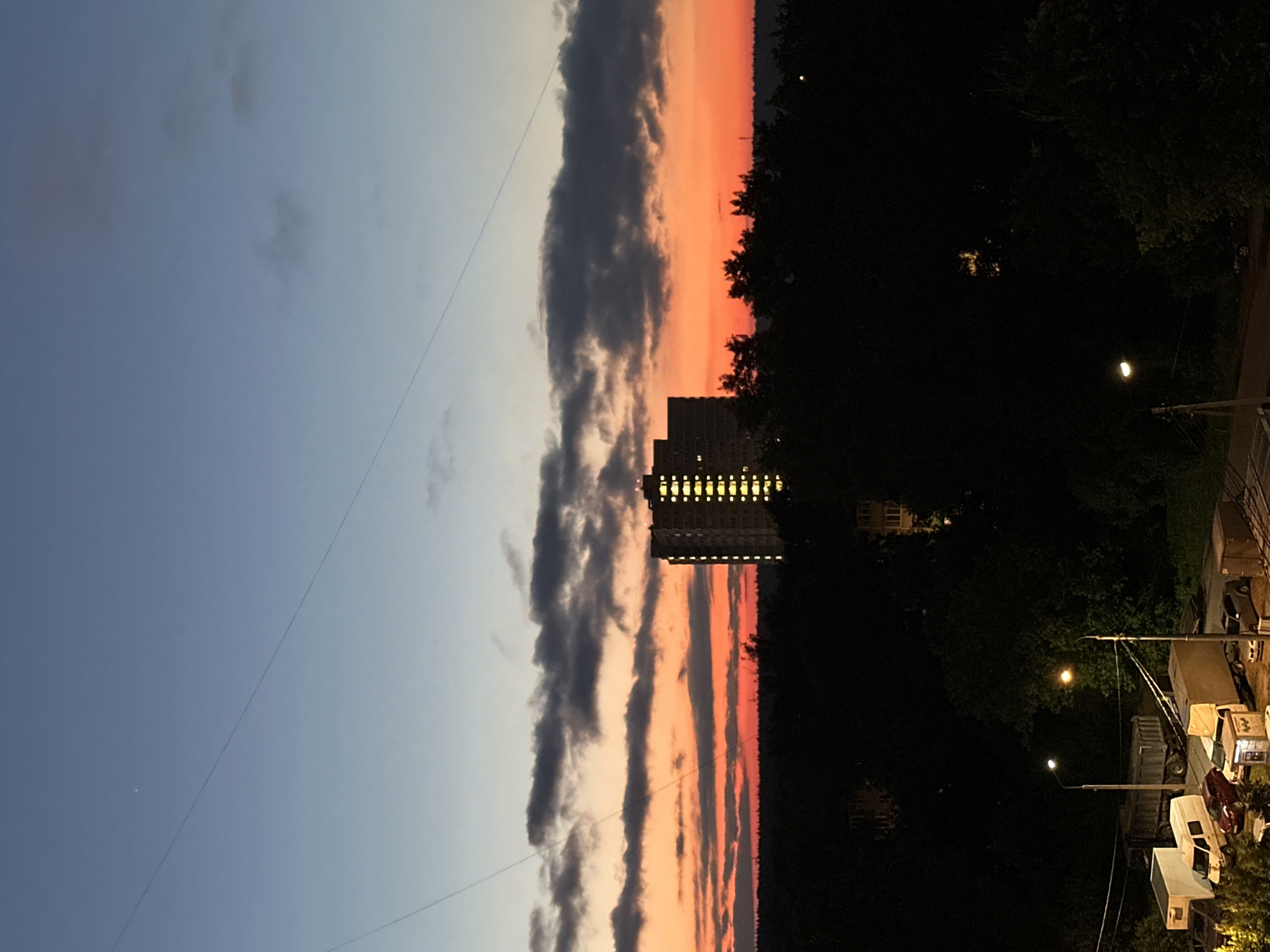 The image depicts a dusky sky over a city at dusk. A large, illuminated apartment building stands tall amidst a dense forest of trees, its windows glowing with interior lights. The sky is a gradient of blue at the top, transitioning to vibrant orange and red hues near the horizon, streaked with dark clouds. In the foreground, a street scene is visible, with parked vehicles including vans, trucks, and cars, illuminated by streetlights. The overall impression is one of urban life settling in for the evening, with the natural landscape of trees providing a dark, silhouetted backdrop.