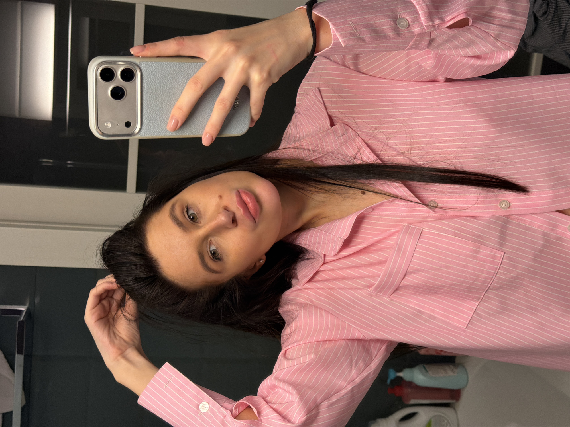 An indoor mirror selfie features a young woman looking directly at the camera, with the image rotated 90 degrees counter-clockwise from a standard portrait orientation. Her head appears towards the bottom left of the frame. She holds a light blue-gray smartphone with a matching case and three camera lenses in her right hand, which is positioned high in the frame.
The woman has long, dark hair that cascades around her shoulders. Her eyes are a light brown or hazel, and her lips are slightly parted in a subtle, neutral expression. She wears a pink button-up shirt with thin white vertical stripes, featuring a collar and a single breast pocket on the left side. A black elastic band is visible on her right wrist, and her fingernails are painted with light pink polish.
The background, visible through the mirror's reflection, consists of dark, reflective panels or tiles, suggesting a modern bathroom or hallway setting in Khimki, Russia. Various household items, including a light blue, a red, and a white bottle (possibly cleaning products or toiletries), are partially reflected on a surface in the bottom right corner of the image. The lighting is bright and even, illuminating the scene clearly. No discernible text is visible in the frame.