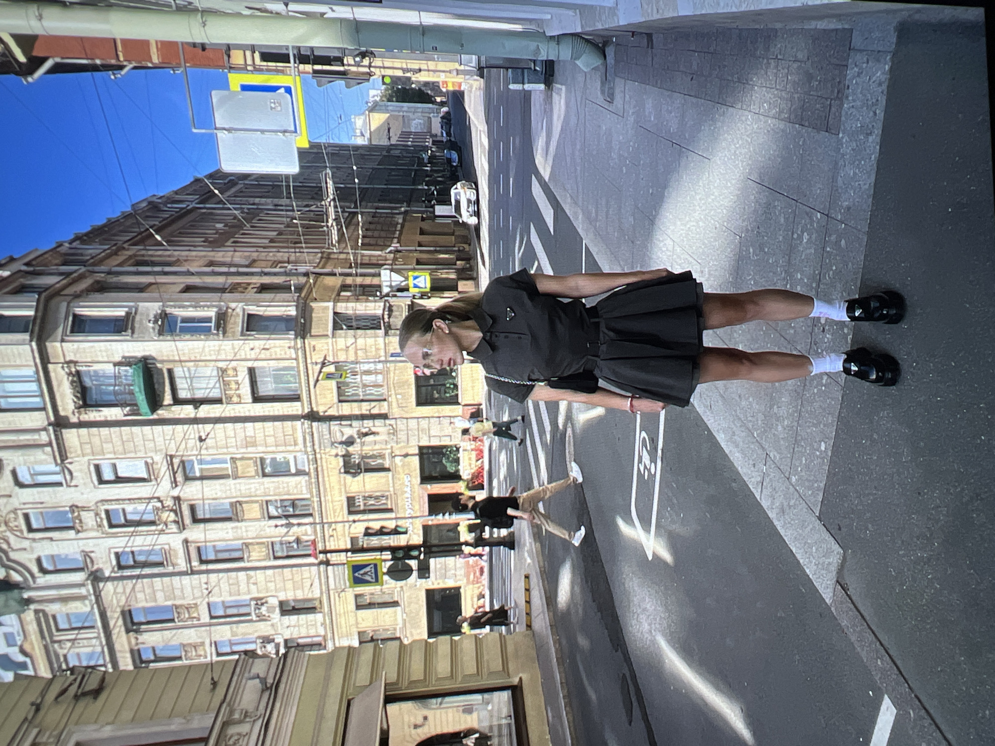 A young woman stands on a sidewalk in Saint Petersburg, Russia, facing right. She is wearing a dark collared dress, white socks, and black platform shoes. Her hair is pulled back in a ponytail, and she wears glasses. In the background, several people are walking across a street. Buildings with ornate architecture line the street. A street sign is visible on the left side of the image. The sky is clear and blue.