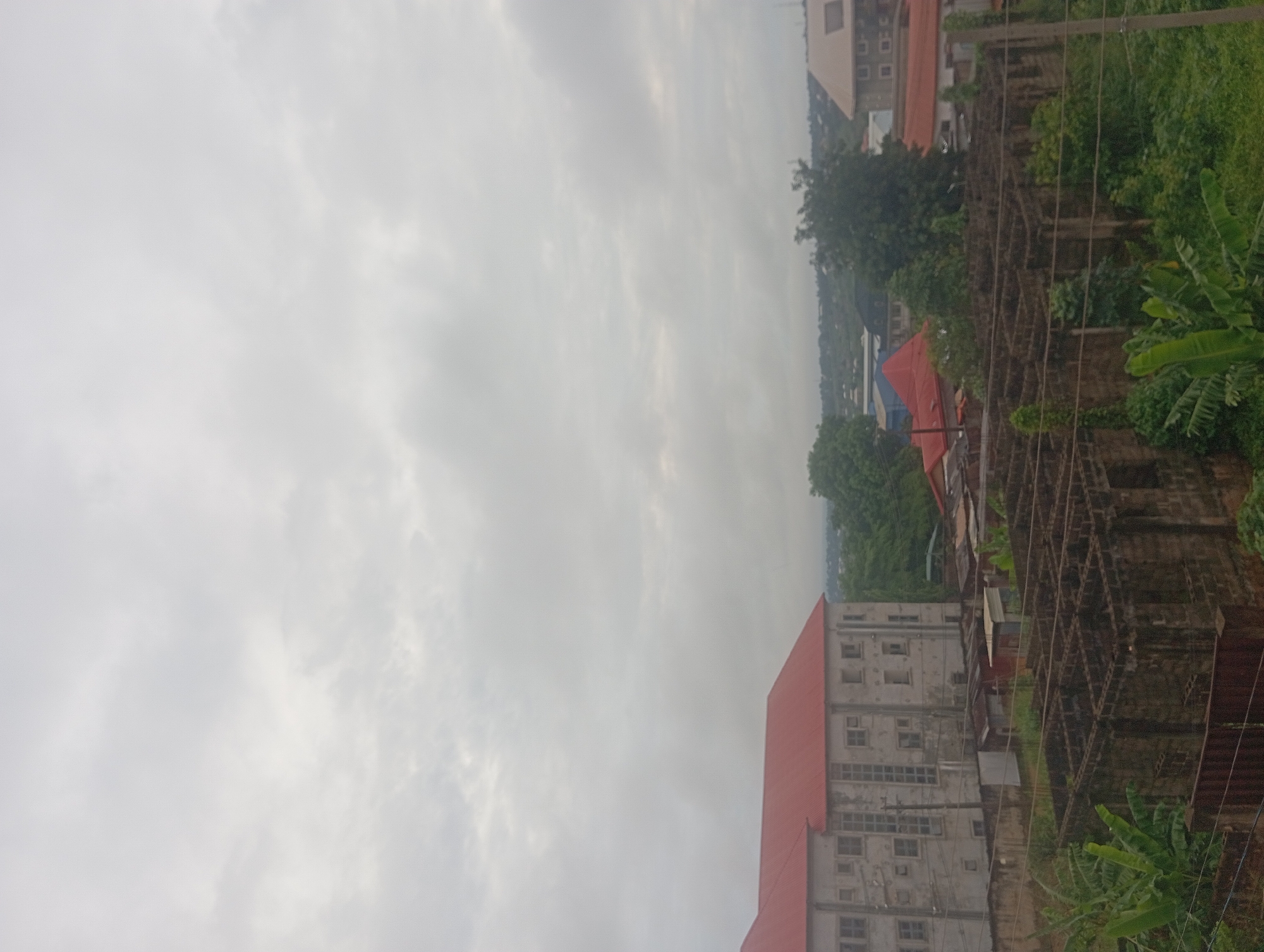 The image shows a view in Nkpor, Nigeria. The sky is overcast with a dense cloud cover. A tall, concrete building with a red roof is visible in the foreground. Adjacent to it is a dilapidated structure, possibly a construction site or ruins, overgrown with vegetation. Further in the distance are other buildings, trees, and a glimpse of the urban landscape. Banana trees are present in the lower right corner. Electrical wires are strung across the foreground.