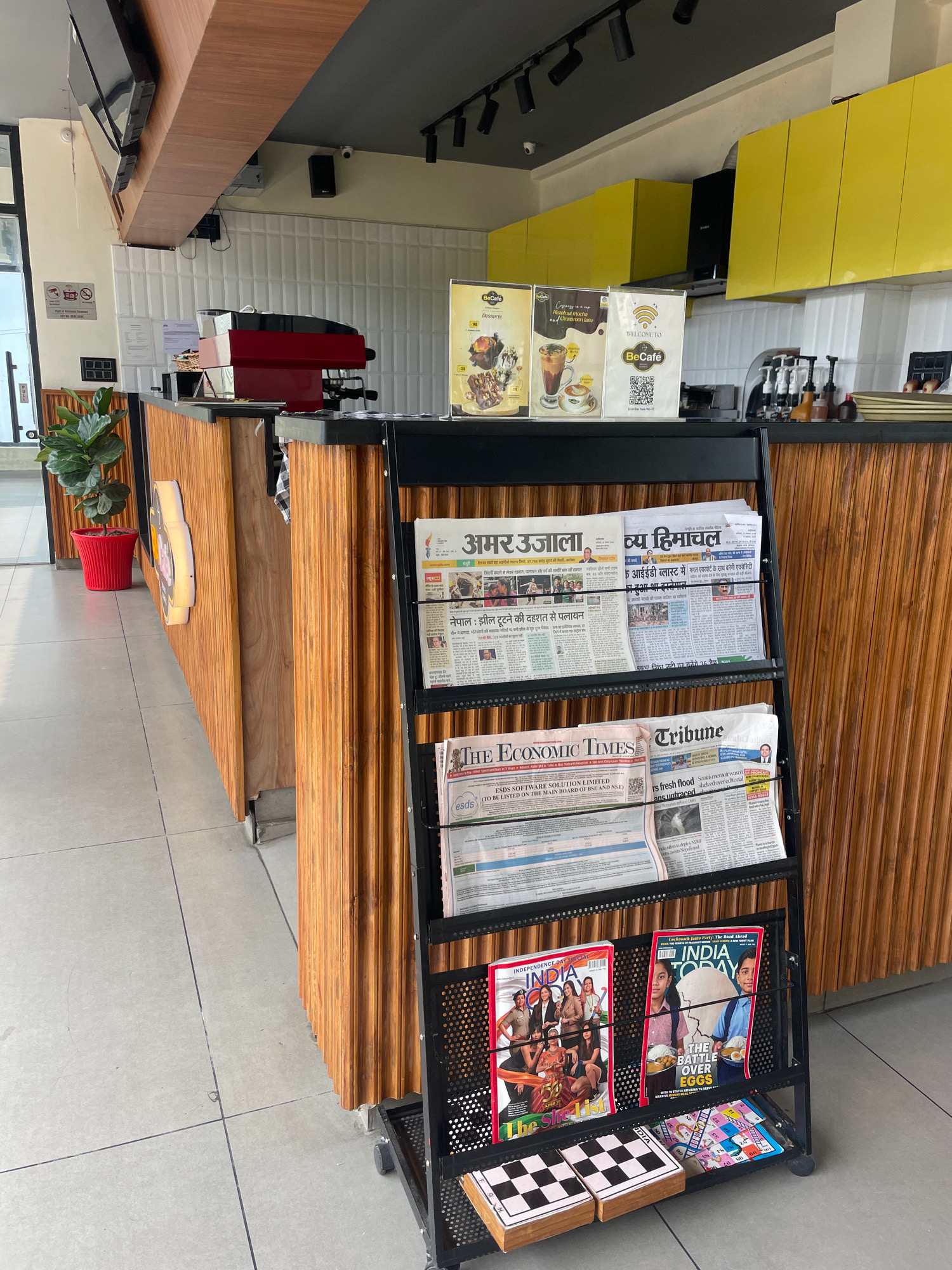 The image captures the interior of a cafe with a modern design. A prominent feature is a black metal rack displaying various newspapers and magazines. The newspapers appear to be in Hindi, with visible titles like "अमर उजाला" and "व्य हिमाचल." The magazines include "India Today" and "The Size List." In the background, a wooden counter is visible, with a red espresso machine and shelves stocked with bottles. Yellow cabinets are mounted on the wall above the counter, suggesting a vibrant and contemporary aesthetic. A potted plant in a red pot adds a touch of greenery to the scene. The floor is tiled in a light grey color, and the overall environment suggests a well-maintained and inviting space for customers to relax and read. The presence of newspapers and magazines indicates that this is likely a public establishment where patrons can access reading materials.