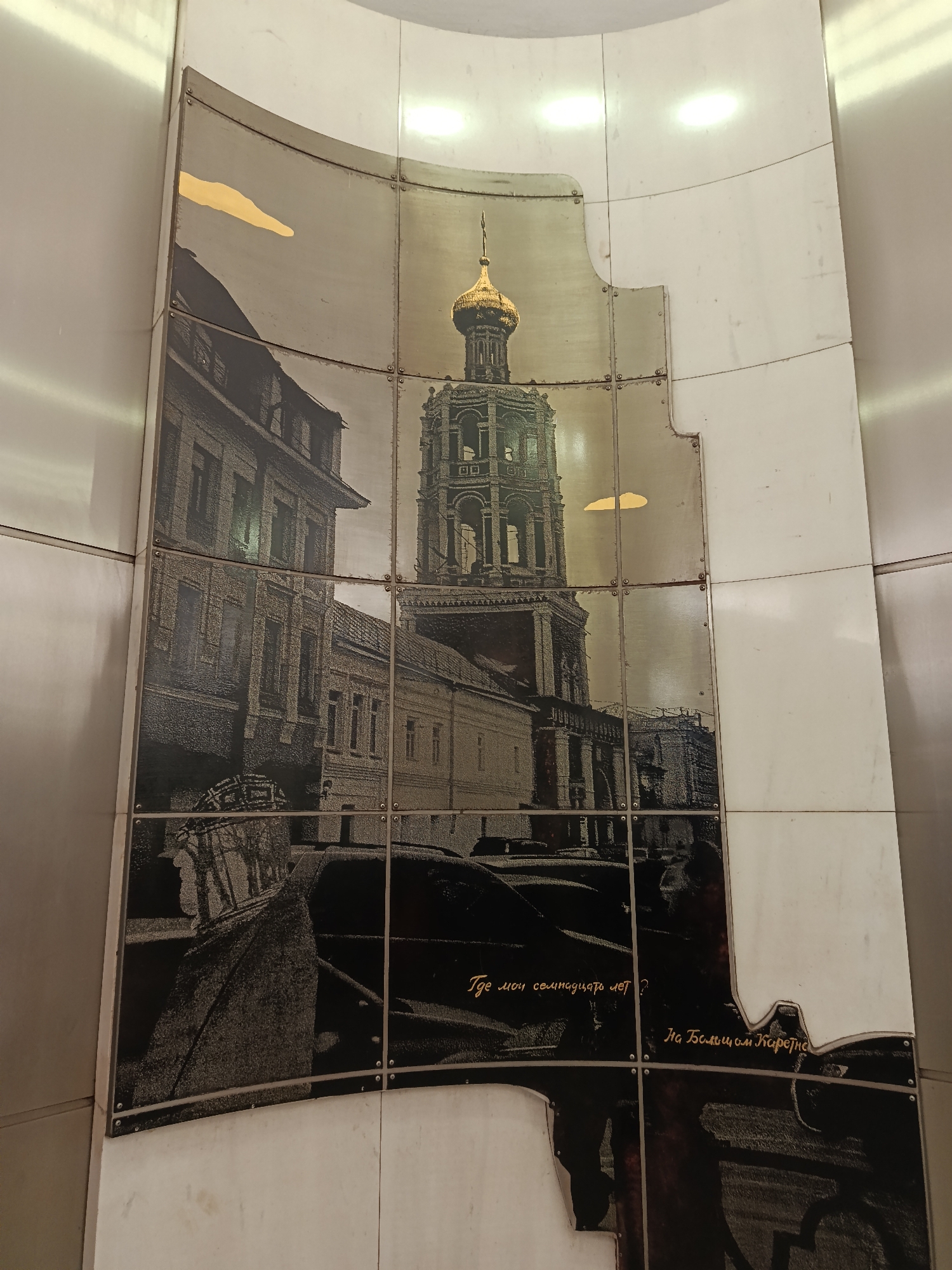 A large, multi-panel mural is displayed on a curved, light-colored wall, likely within an indoor public space. The artwork features a monochromatic cityscape, rendered in tones of gray or sepia, with distinct metallic gold highlights.
The central subject of the mural is a tall, ornate bell tower crowned with a prominent golden dome. Surrounding buildings, appearing to be older architectural styles, are partially visible on either side of the tower. The lower portion of the image shows dark, indistinct shapes resembling parked cars and possibly blurred human figures, silhouetted against the foreground. The sky area above the buildings includes two abstract, cloud-like shapes, also rendered in metallic gold.
The mural is constructed from multiple rectangular panels, possibly metal or ceramic, with visible rivets securing them. These panels form an irregular, cutout shape for the main image against the plain, lighter panels of the surrounding wall. The photographic imagery has a textured, almost grainy quality.
Two lines of text in a script font are visible over the dark, lower section of the mural. The first line reads "Где мои семнадцать лет ?" (Where are my seventeen years?), and the second line reads "На Большом Каретном" (On Bolshoy Karetny).