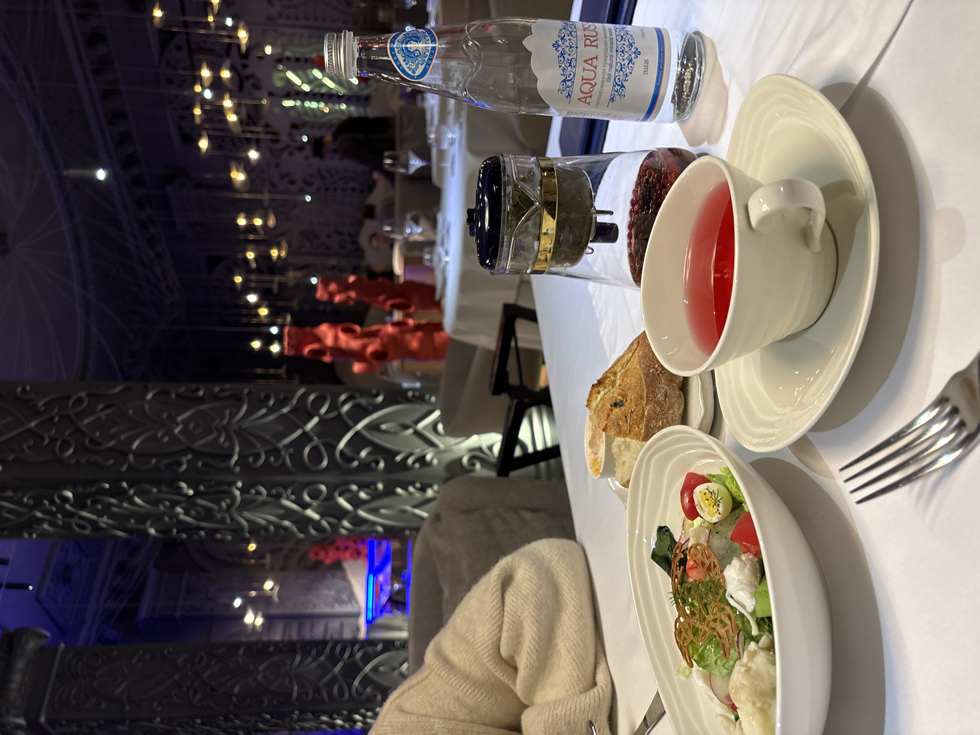 The image depicts an indoor dining scene at Trattoria Formaggi, located at Ulitsa Karla Marksa, 149, Krasnoyarsk, Russia. The perspective is a close-up, slightly elevated view of a table set for a meal.
In the immediate foreground, a white tablecloth covers the table. A silver four-tined fork rests on the tablecloth, pointing towards the upper left. A white oval bowl contains a salad composed of green lettuce, halved quail eggs, cherry tomatoes, radish slices, a creamy white cheese or dressing, and a delicate, crispy golden-brown garnish. Adjacent to the salad bowl, a small white plate holds slices of rustic bread and a ramekin containing a light-colored spread.
To the right of the food, a clear glass bottle of "AQUA RUS" still natural mineral water, with a blue and white label indicating an expiration date of "19.12.25," stands upright. Next to the bottle is a clear glass infuser with a dark lid, containing red berries (likely cranberries or lingonberries) at the bottom and a mesh basket filled with tea leaves above. A white ceramic teacup, holding a reddish-pink beverage, sits on a matching saucer.
On the far left, the arm and torso of a person wearing a light-colored, possibly cream-colored or beige, knitted sweater are partially visible, indicating they are seated at the table.
The background reveals the restaurant's interior. Dominating the decor are large, dark, cylindrical columns adorned with intricate, swirling, ornate patterns. The ceiling is dark, featuring numerous small, warm-toned pendant lights suspended at varying heights, creating a shimmering effect. In the mid-ground, blurred red sculptural elements, possibly large vases or abstract art, are visible between the columns. Several other white-clothed tables with chairs are discernible in the distance, some occupied by blurred figures. A figure in a dark garment is partially visible behind the Aqua Rus bottle. Areas of blue ambient lighting are present further into the background, hinting at additional decorative elements or a bar area. The overall lighting is dim, atmospheric, and focused, highlighting the tables and decorative features.