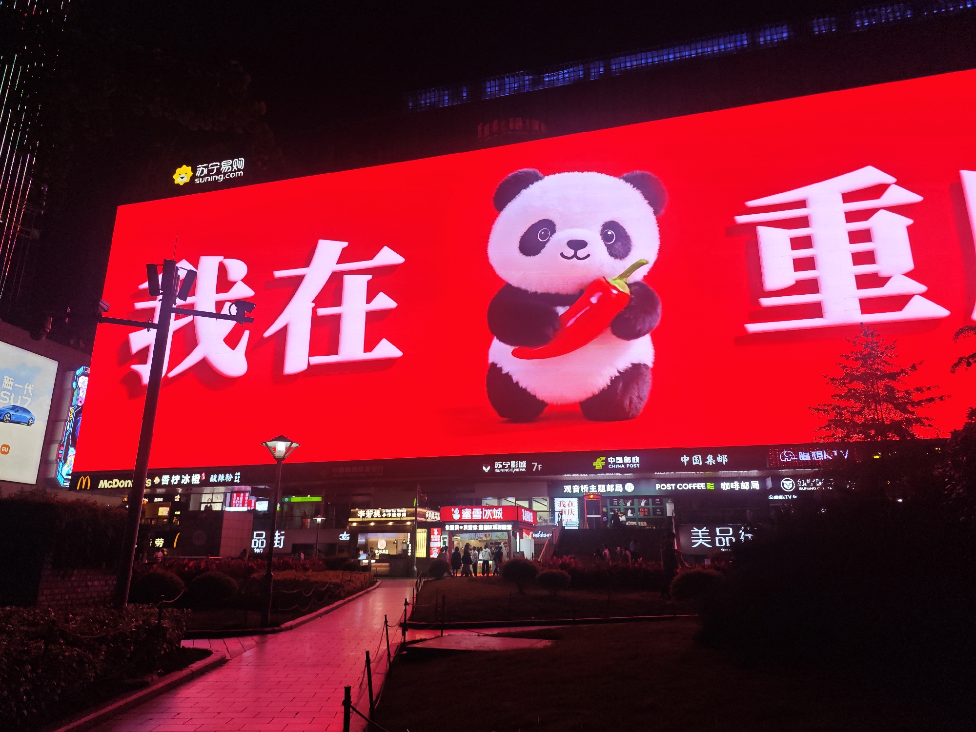 This nighttime photograph showcases a bustling commercial streetscape in Liangjiang, China, illuminated by numerous signs and a dominant LED screen. The main subject is a massive LED billboard covering the facade of a building, displaying a vibrant red background. On this screen, large white Chinese characters "我在" (Wǒ zài, meaning "I am in" or "I am at") are prominently featured alongside a playful cartoon panda holding a bright red chili pepper. To the right of the panda, the character "重" (Zhòng) is visible, suggesting the full phrase "我在重庆" (I am in Chongqing), which aligns with the provided location context of Liangjiang (a district of Chongqing). Above the main display, the logo and text for "苏宁易购 suning.com" are displayed.
Below the large LED screen, a variety of businesses line the street level, their storefronts and signs brightly lit. Visible establishments include a McDonald's, "苏宁影城 7F" (Suning Cinema on the 7th floor), "中国邮政 CHINA POST" (China Post), "观音桥主题邮局" (Guanyinqiao Theme Post Office), and "POST COFFEE 咖啡邮局" (Post Coffee). Other shops like "蜜雪冰城" (Mixue Bingcheng), "唱想KTV," and "美品社" are also visible. On the far left, a narrower vertical LED screen advertises "新一代 SU7" (New Generation SU7) with a blue car image and the Xiaomi logo.
The ground level features paved pathways, landscaped areas with bushes and small trees, and streetlights providing additional illumination. The intense red light from the main LED screen casts a reddish glow across the entire scene, particularly on the walkways and nearby foliage. Several people can be seen walking on the pathways and near the shops, contributing to the lively urban atmosphere. A small, illuminated decorative tree is visible on the ground level near the bottom right of the main screen. The time of day is clearly night, indicated by the dark sky and pervasive artificial lighting.