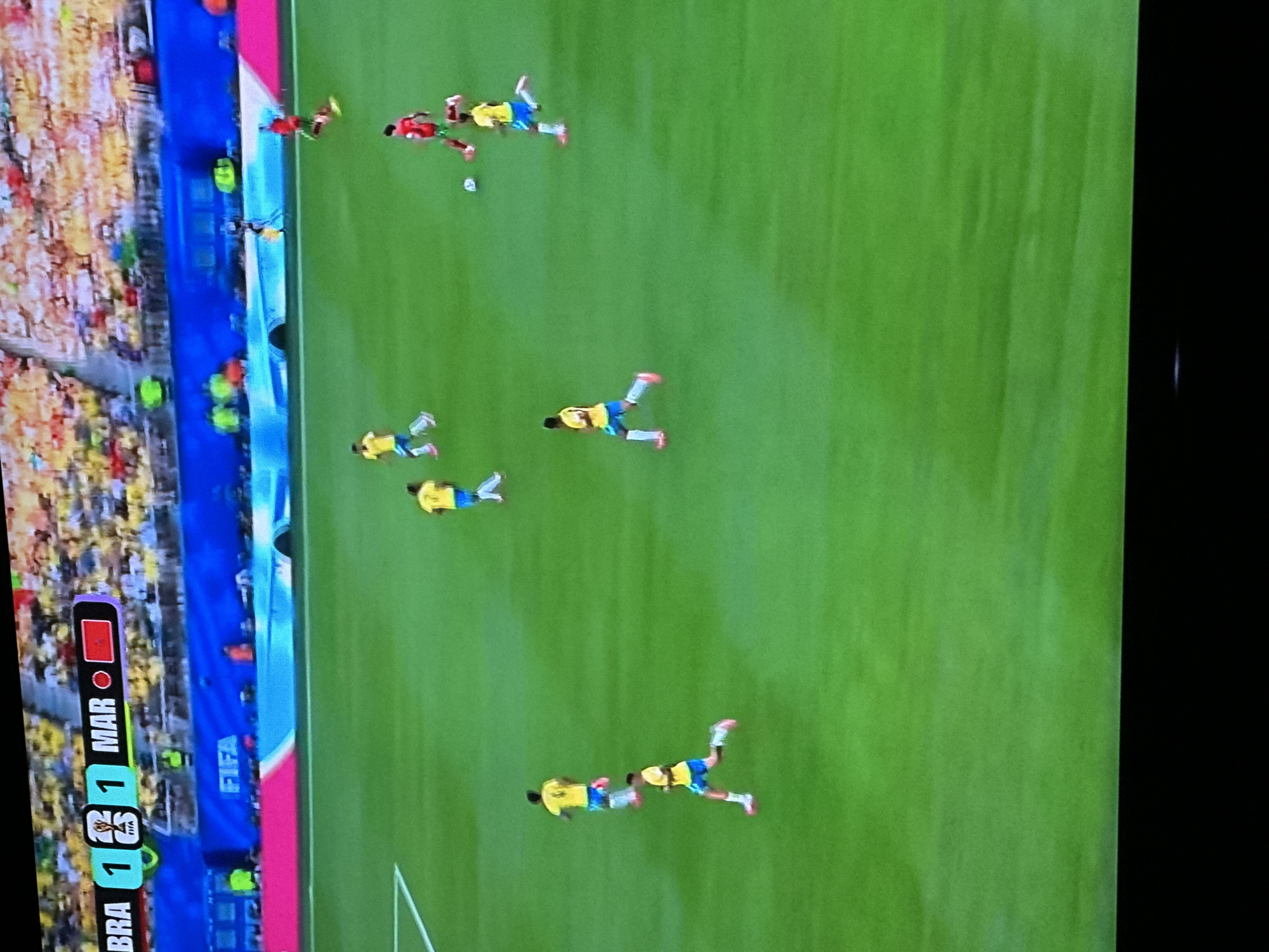 The image captures a soccer match in progress on a vibrant green field. In the upper left corner, a scoreboard displays "BRA 1 2 1 MAR 🔴 🇲🇦". Several players in yellow jerseys with blue shorts are visible on the field, running and positioned across the pitch. One player in a red jersey is also present. The background reveals a stadium filled with spectators wearing a mix of yellow and white attire. A blue barrier with the "FIFA" logo is situated behind the field. The overall scene is characteristic of an outdoor sporting event. The image does not provide any information about the city of Gura Top, Nigeria.