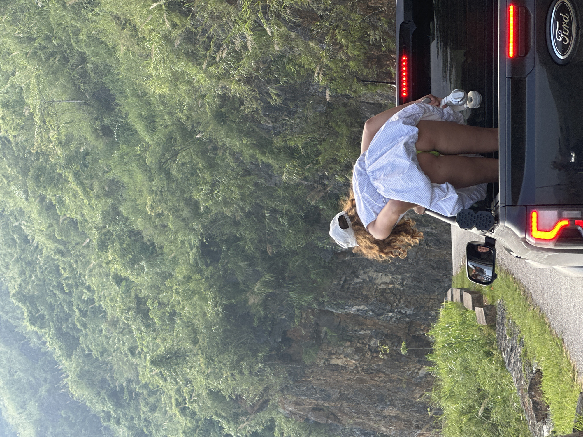 A person wearing a white and blue dress and a cap is bending into the open rear of a dark-colored Ford SUV. The SUV is parked on a road next to a steep, green, vegetated slope. The location is in the Du Già Commune, Vietnam.