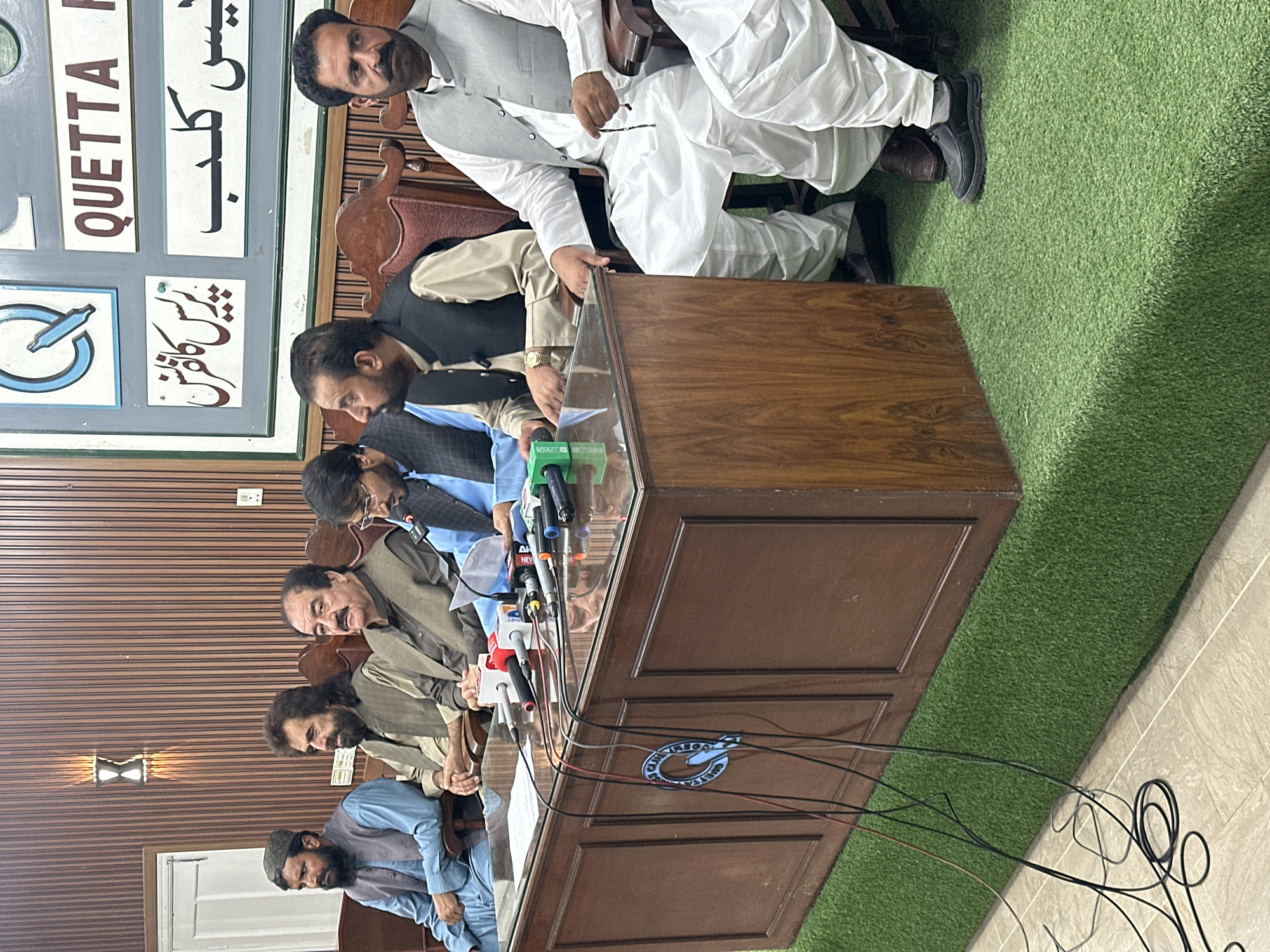 The image depicts a press conference in Quetta City Tehsil, Pakistan, based on the signage in the background that reads "Quetta Press Club" in English and Urdu. Several men are seated behind a large wooden table, facing the camera. Microphones from various news agencies are visible on the table. The setting appears to be indoors, with a faux-grass carpet behind the table and tiled flooring in the foreground. The men are dressed in traditional Pakistani clothing.